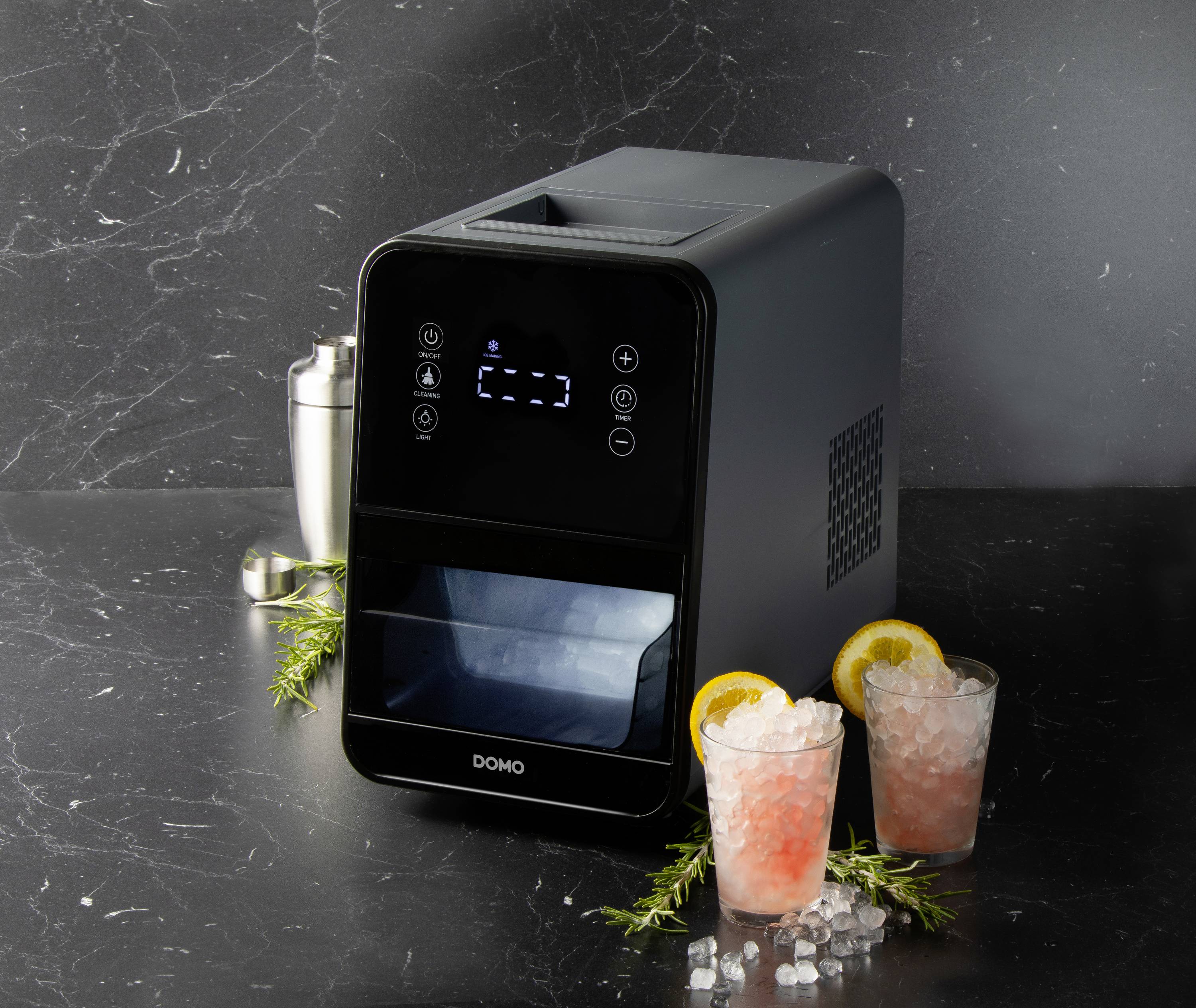 Een zwarte ijsblokjesmachine staat op een tafel met twee glazen roze drank, gegarneerd met citroenschijfjes en kruiden.