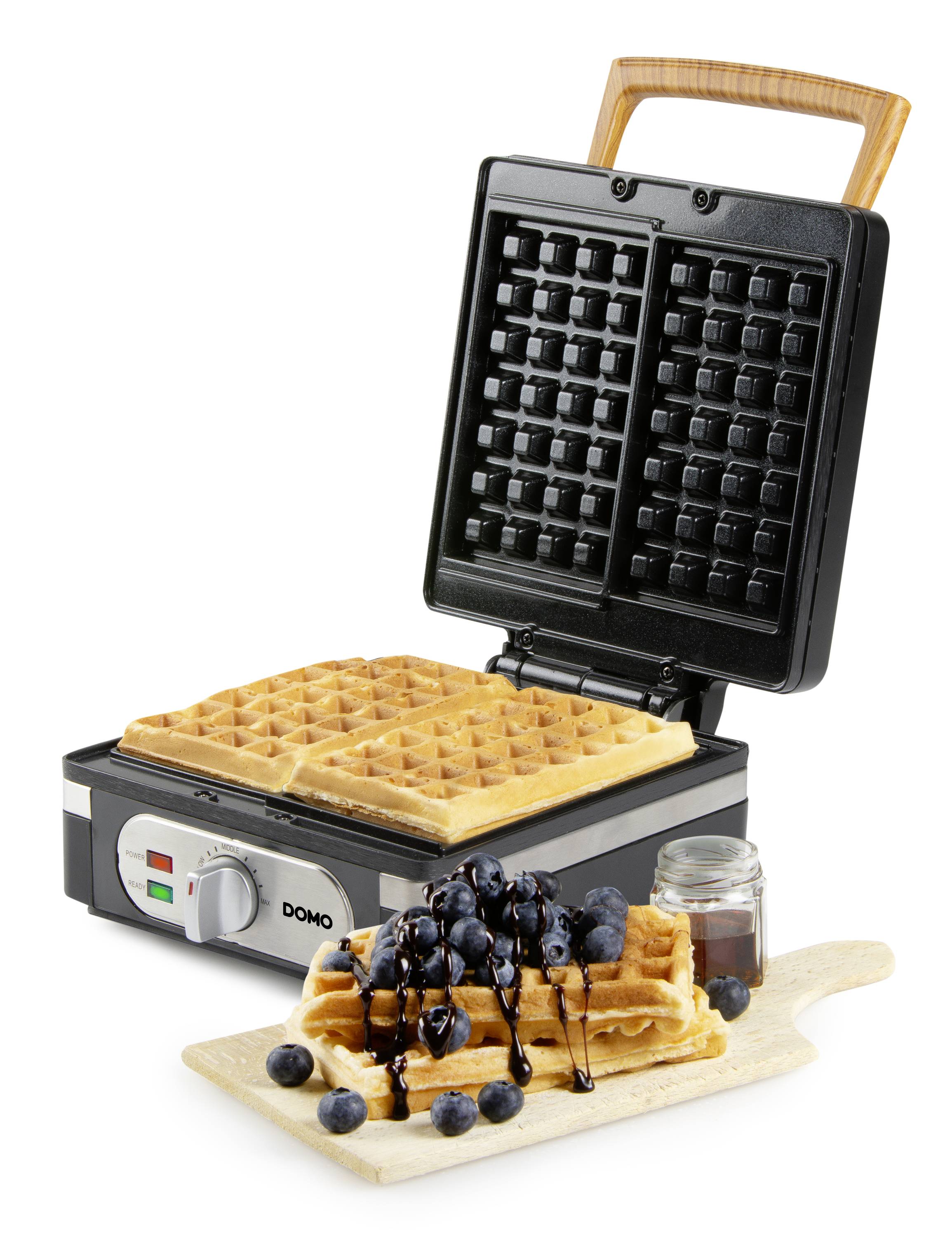 Wafelijzer met twee goudbruine wafels, naast een houten plank met afgewerkte wafels, bosbessen en chocoladesaus.