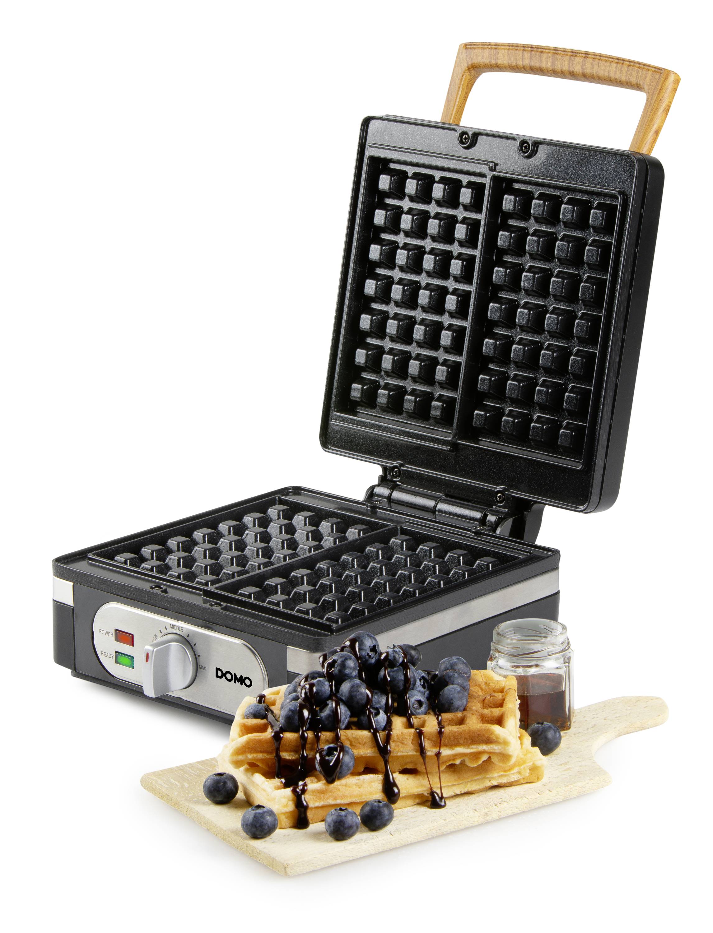 Een wafelijzer met geopend deksel, ernaast een wafel op een houten plank, gegarneerd met bosbessen en chocoladesaus.