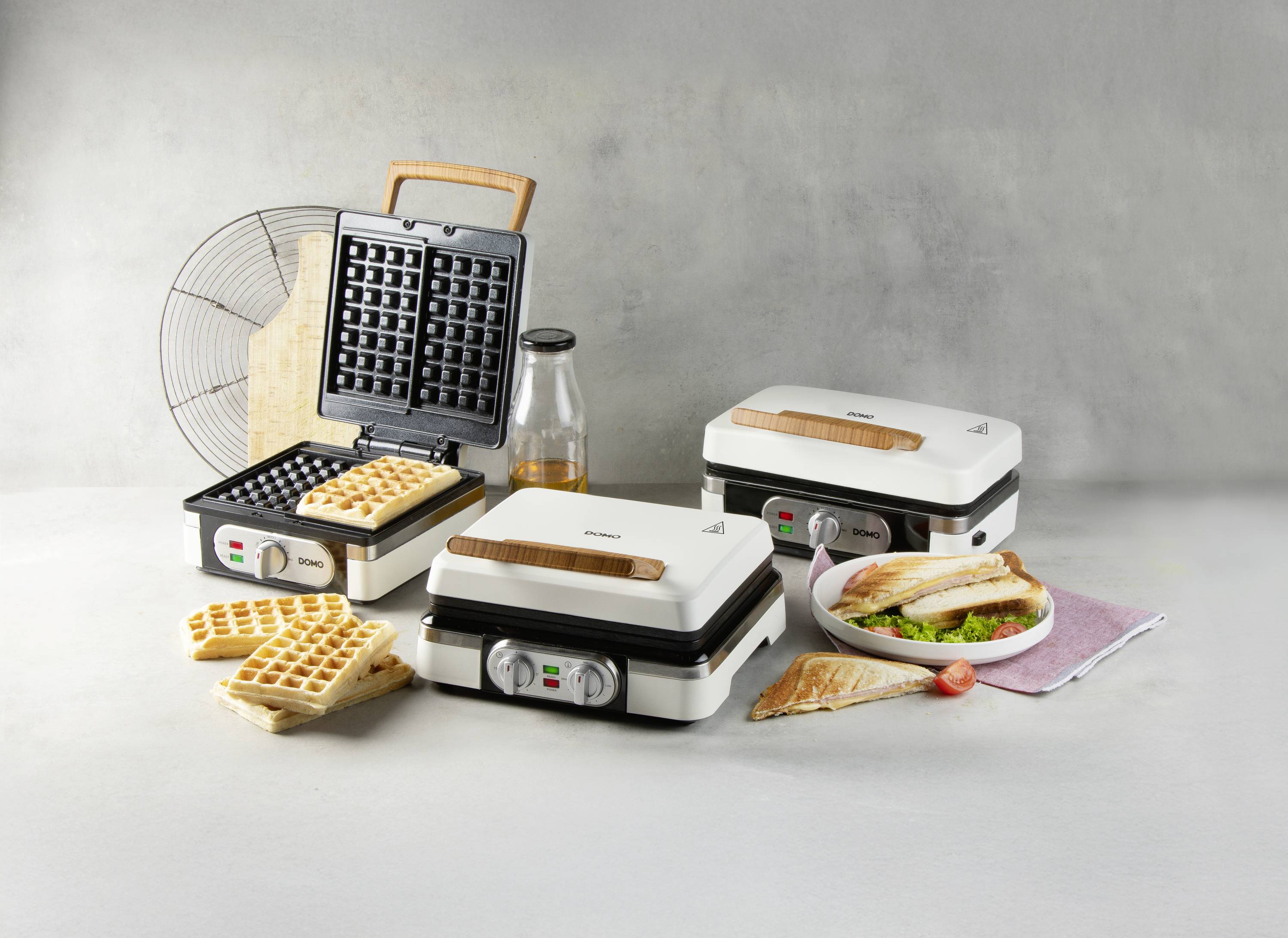 Drie keukengerei op een tafel, elk met verschillende snacks: wafels, sandwiches en gegrilde gerechten. Achtergrond is subtiel grijs.