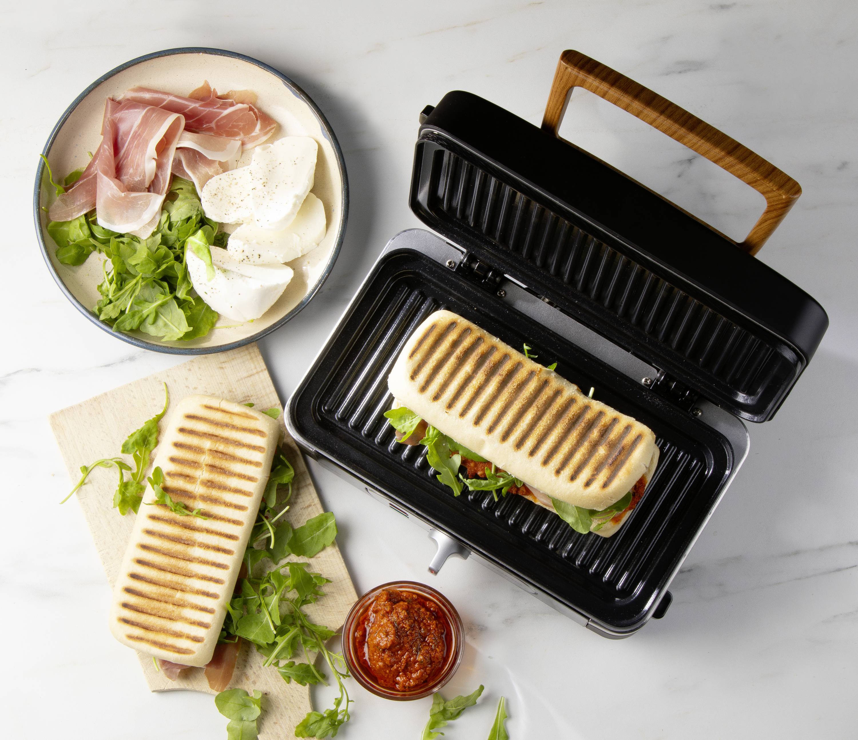 Panini in de sandwichgrill met rucola, ham en mozzarella. Ernaast staat een kom met extra ingrediënten en een glas rode saus.