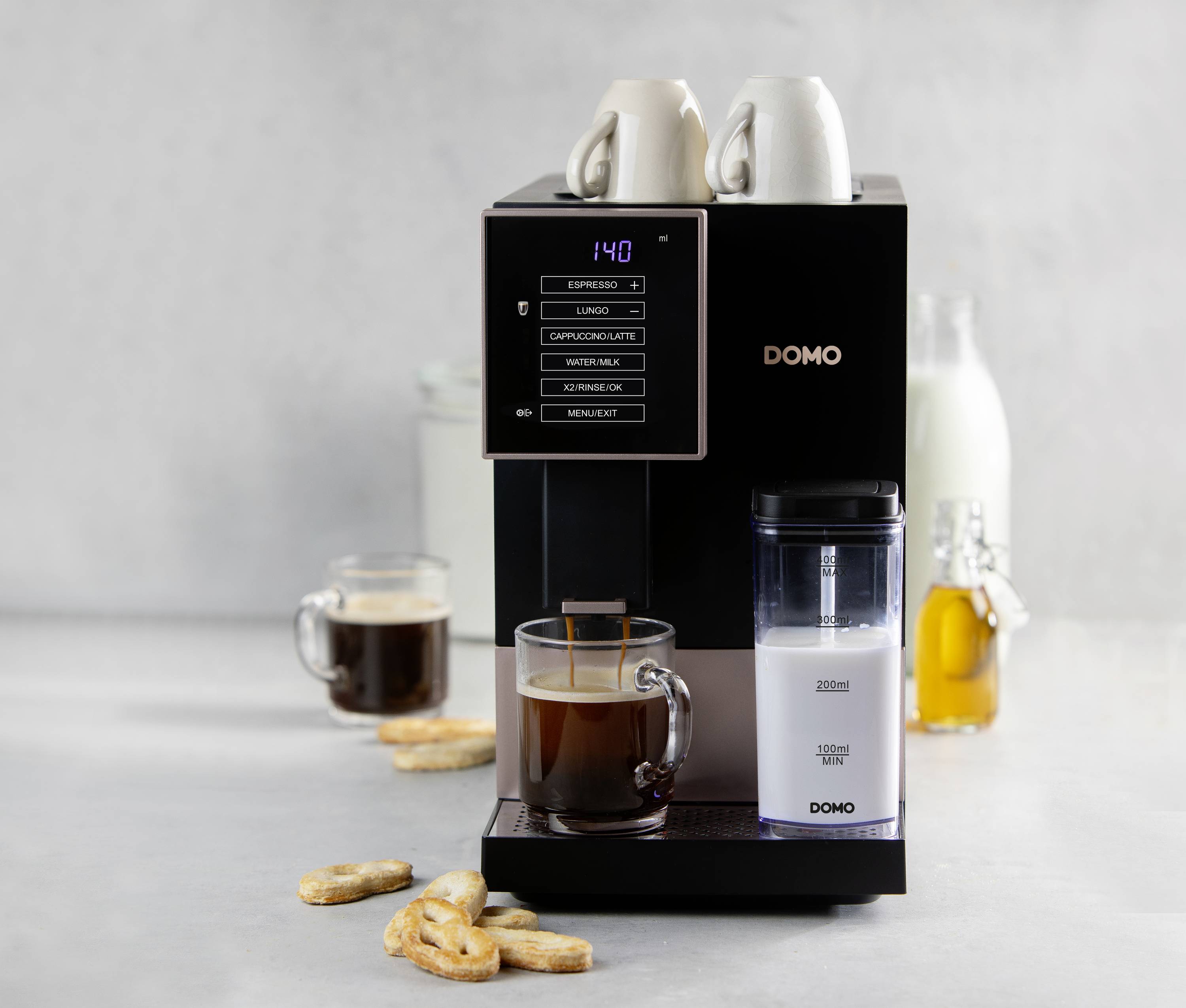 Volautomaat met display toont opties zoals 'Espresso' en 'Cappuccino'. Kopje koffie, koekjes en melkkannetje op de achtergrond.