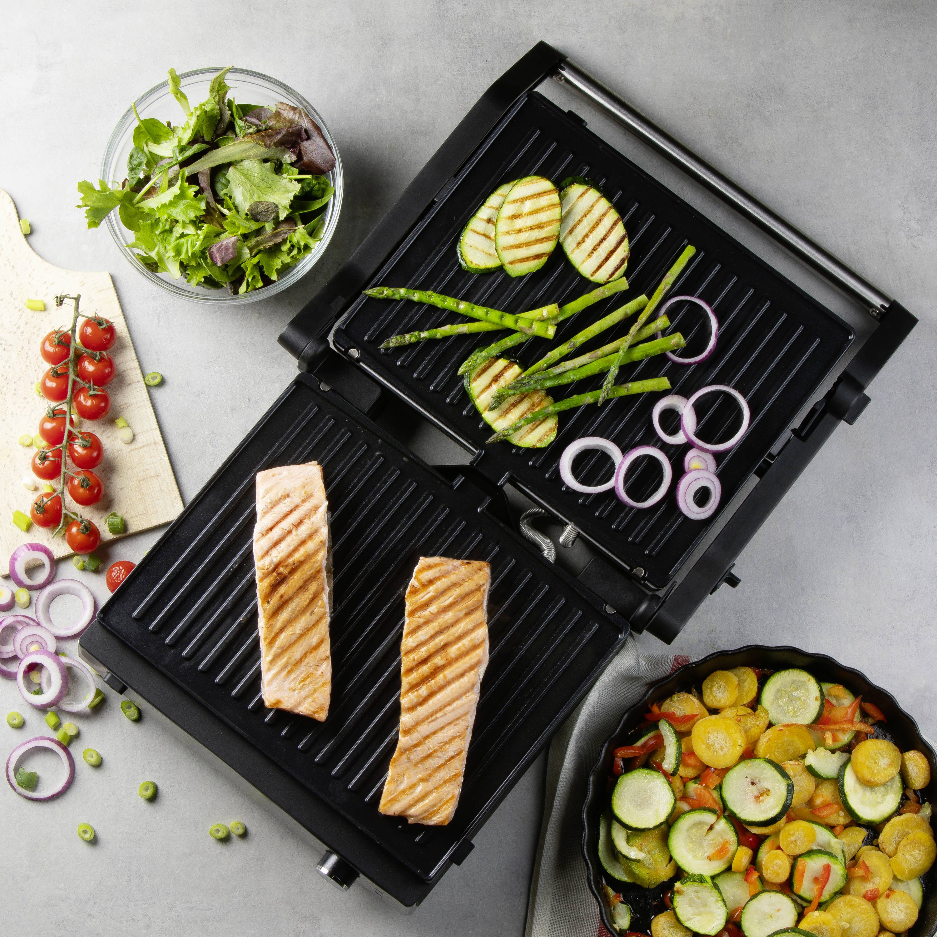 Een elektrische grill met twee zalmfilets bakt, omringd door vers groente zoals courgette, tomaten en sla in kommen en op planken.