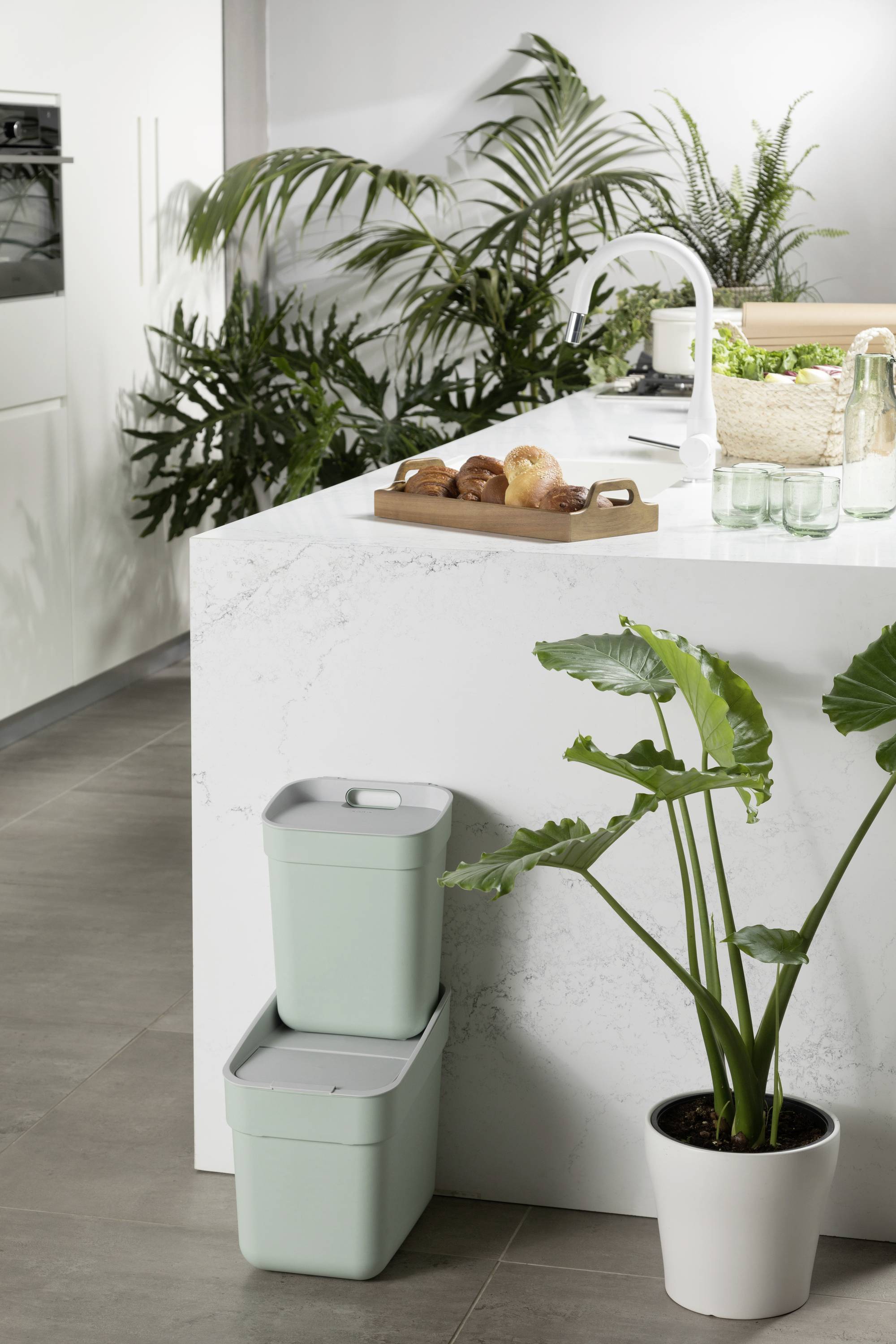 Een moderne keuken met planten, een wit marmeren aanrecht, twee gestapelde, muntgroene containers en een bakplaat met gebak.