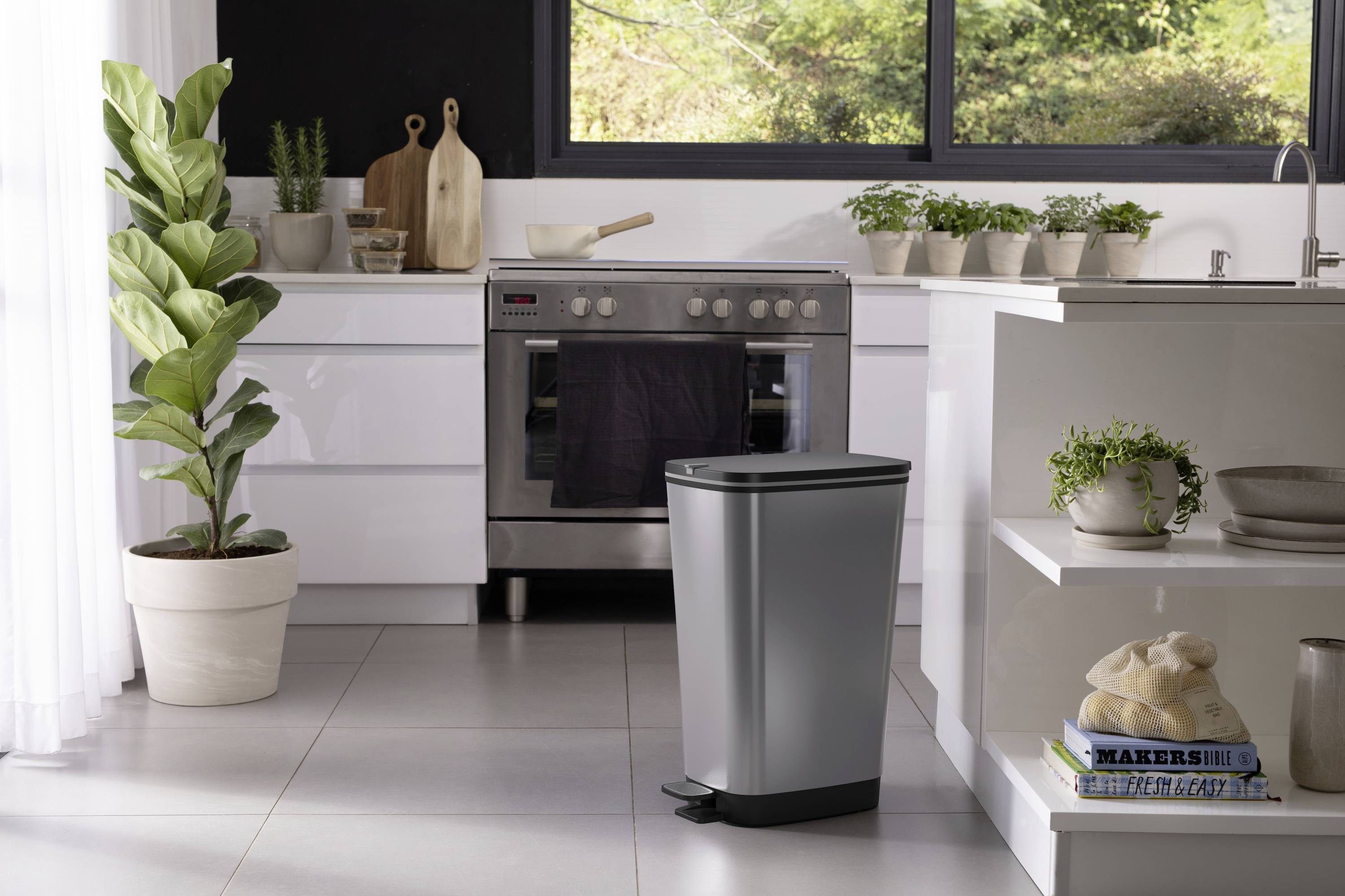 Een moderne keuken met een roestvrijstalen oven, een grijze prullenbak met voetpedaal, planten en een raam met uitzicht op groene natuur.