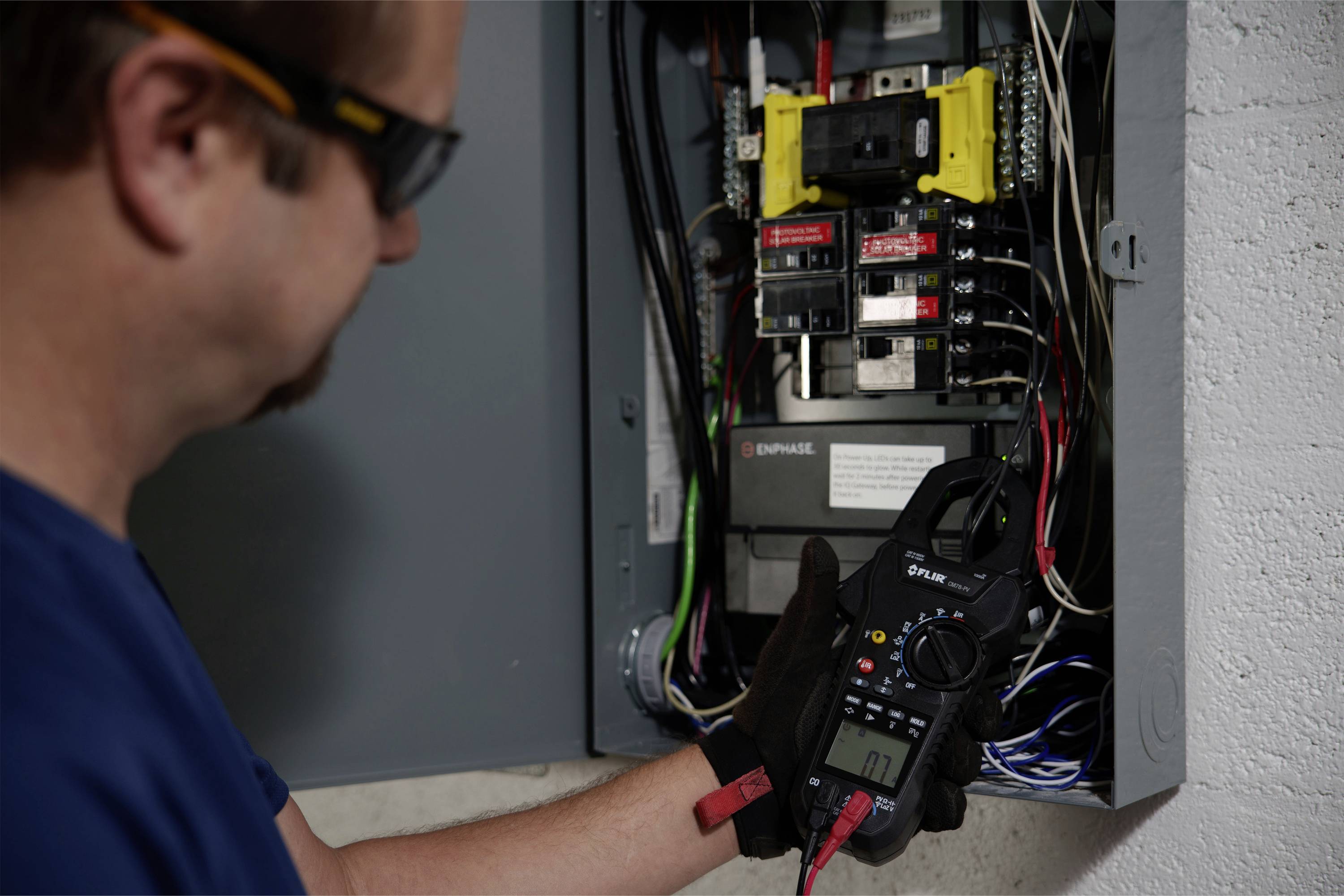 Een elektricien meet met een multimeter de spanning in een geopende zekeringkast. Bovenin zijn verschillende schakelaars en bekabeling zichtbaar.