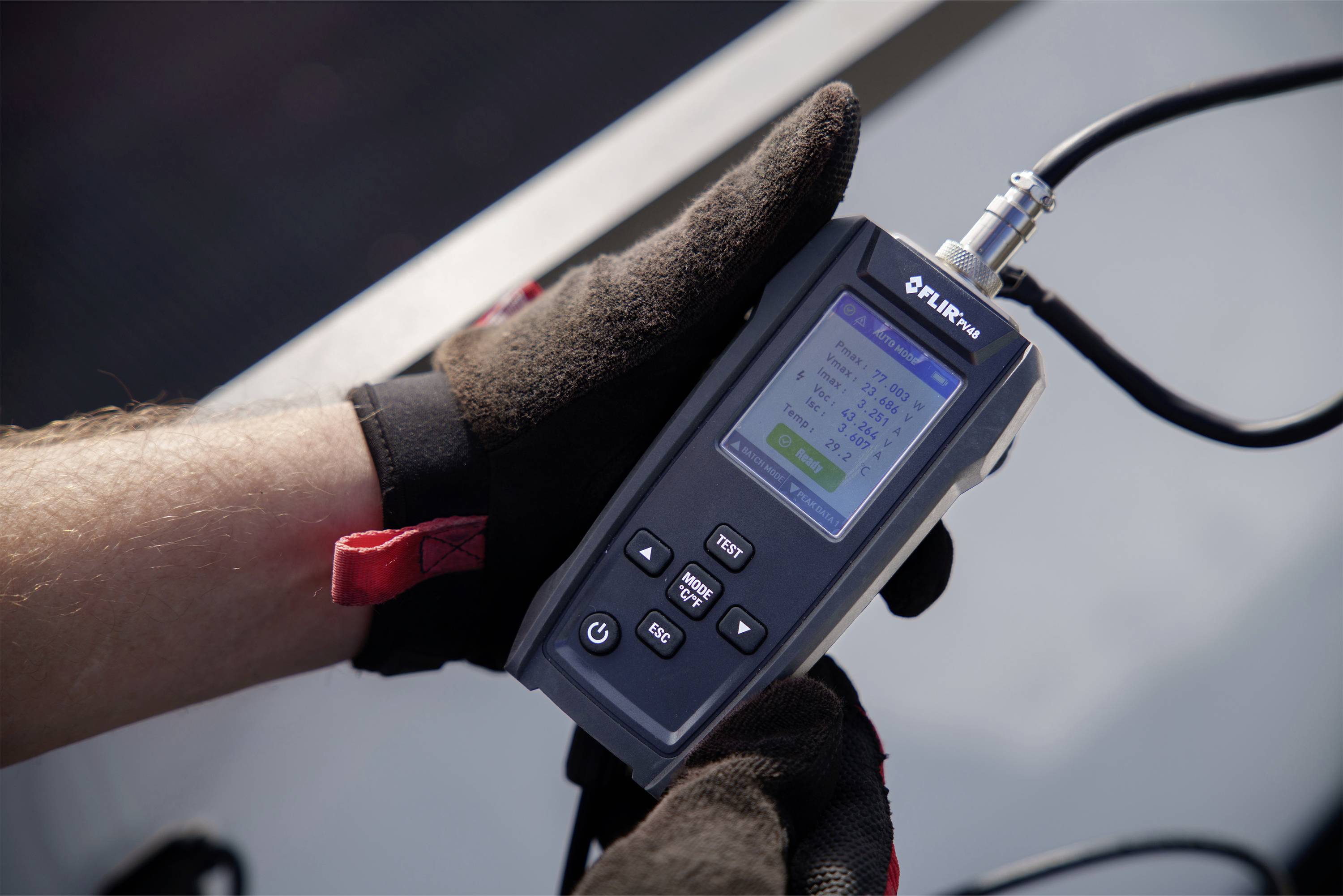 Een persoon houdt een meetapparaat met een display-aanduiding die technische gegevens toont. Het apparaat wordt met beide handen vastgehouden, waarbij de persoon handschoenen draagt.