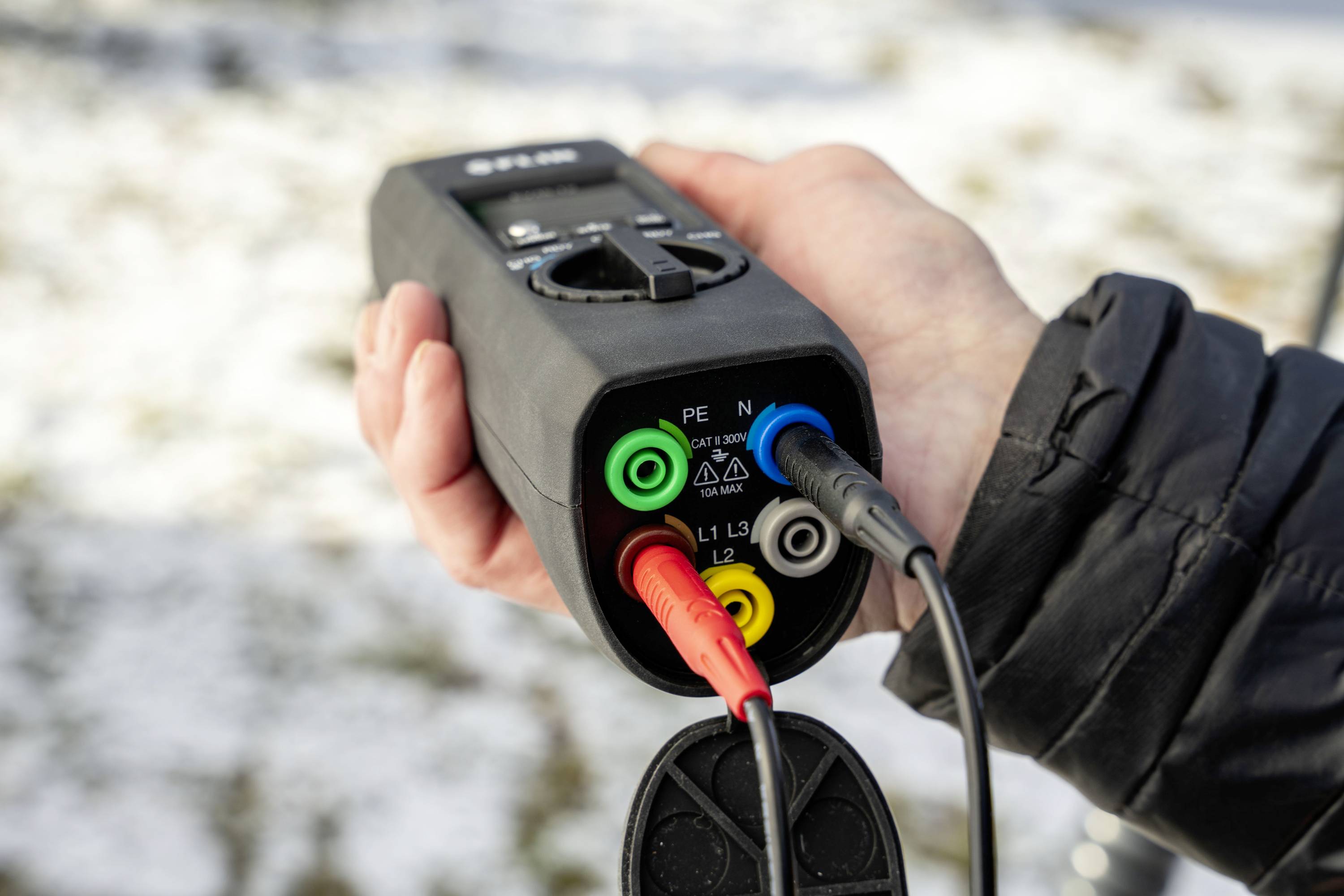 Een persoon houdt een elektrisch testapparaat met kleurrijke kabels, gebruikt voor veiligheids- en functietests van elektrische systemen. Op de achtergrond sneeuw op gras.