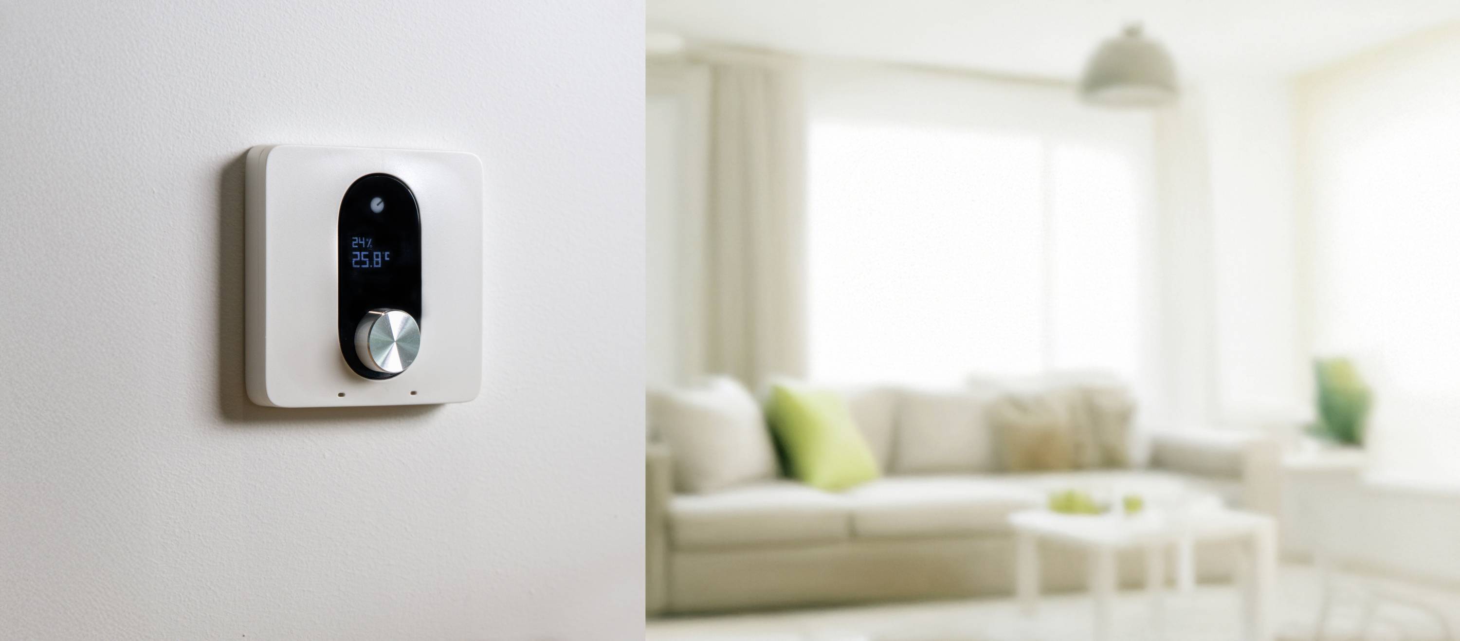 Slimme thermostaat aan witte muur, toont huidige temperatuur. Op de achtergrond wazig een modern woonkamer met bank en salontafel.