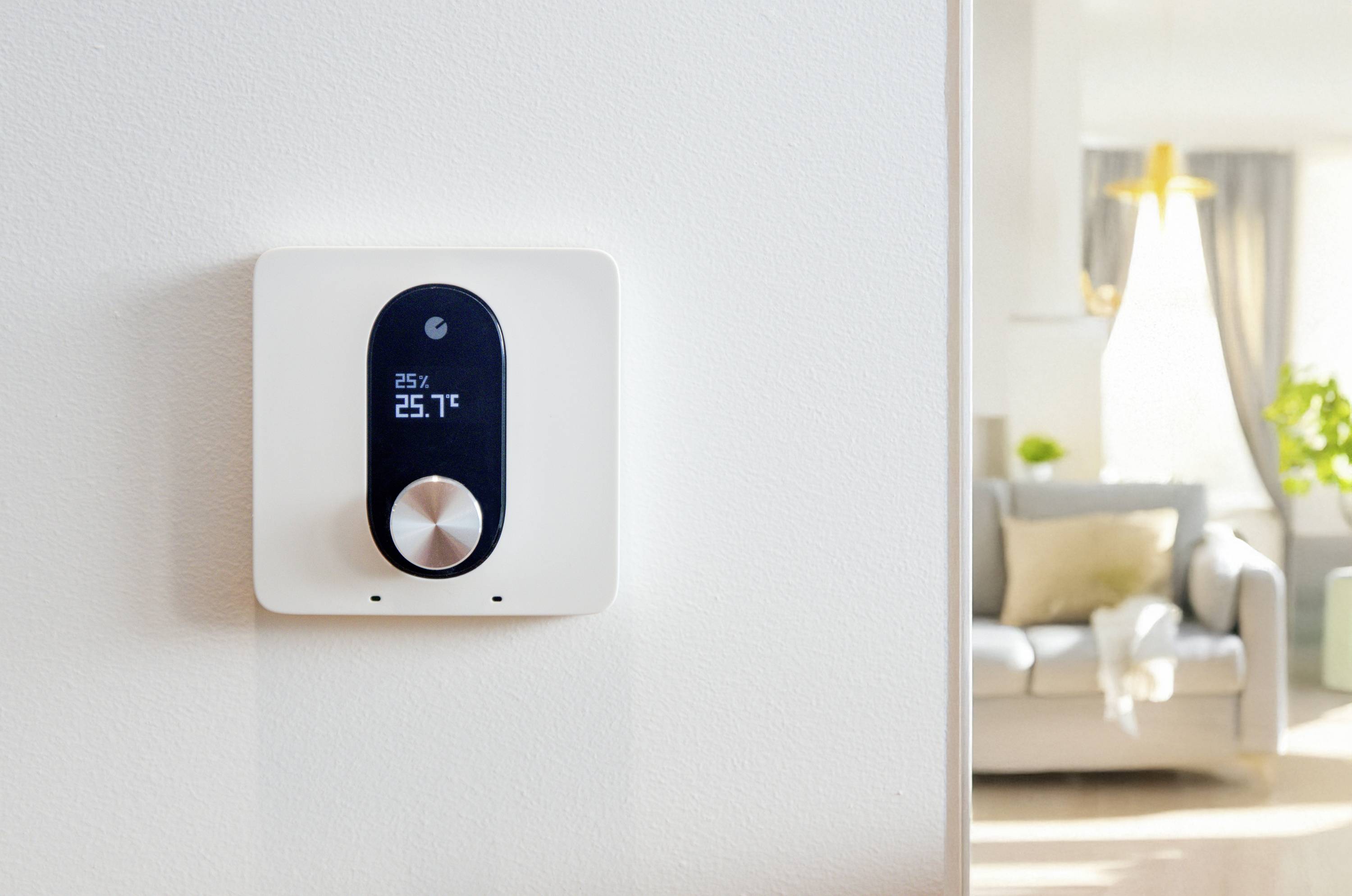 Een modern thermostaat aan de muur toont '25°C, 25,7°C'. Op de achtergrond een gezellige woonkamer met bank en kamerplant.