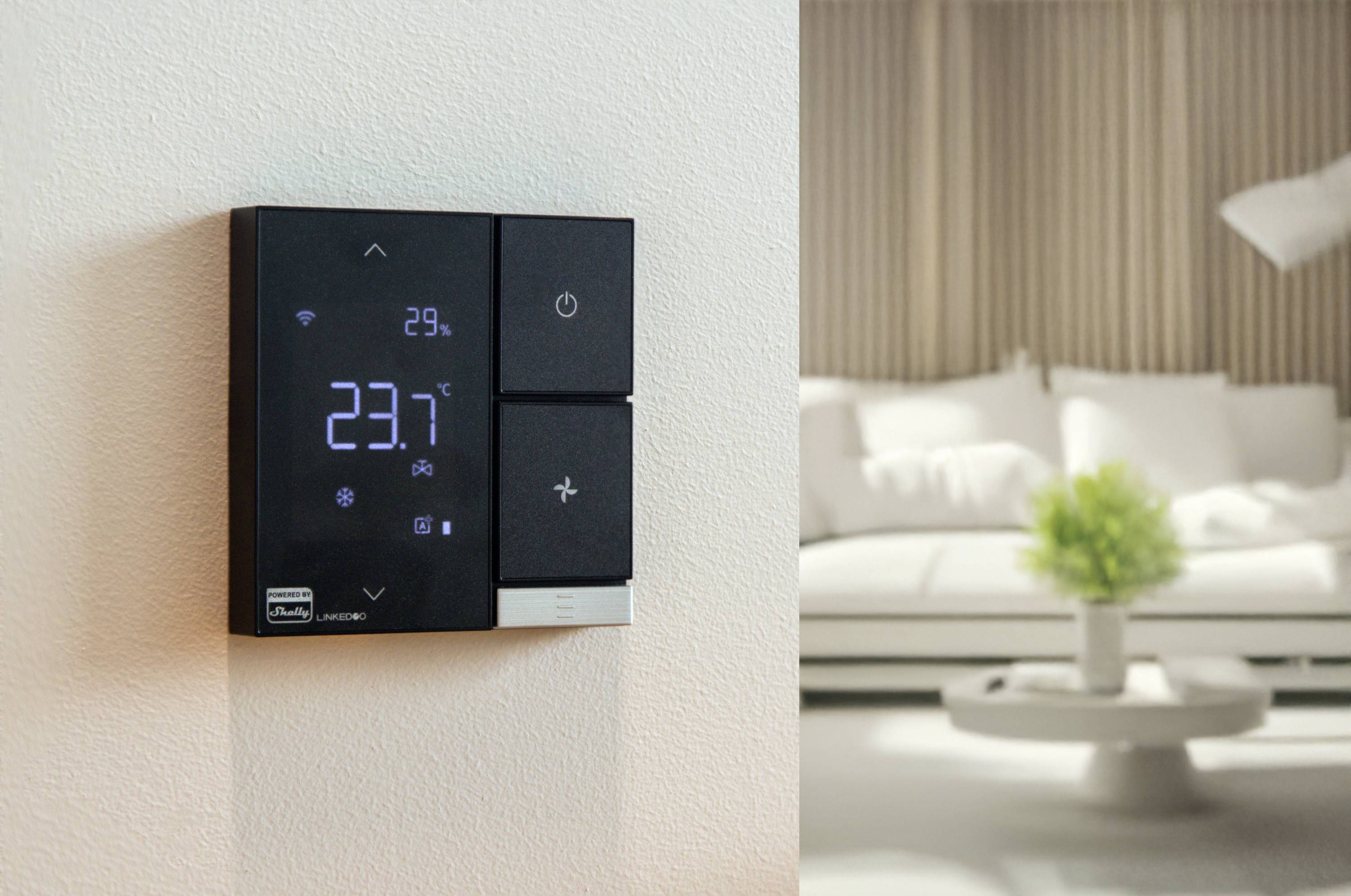 Een intelligent thermostaat aan de muur toont 23,7 graden Celsius. Op de achtergrond is een woonkamer te zien met een witte bank.