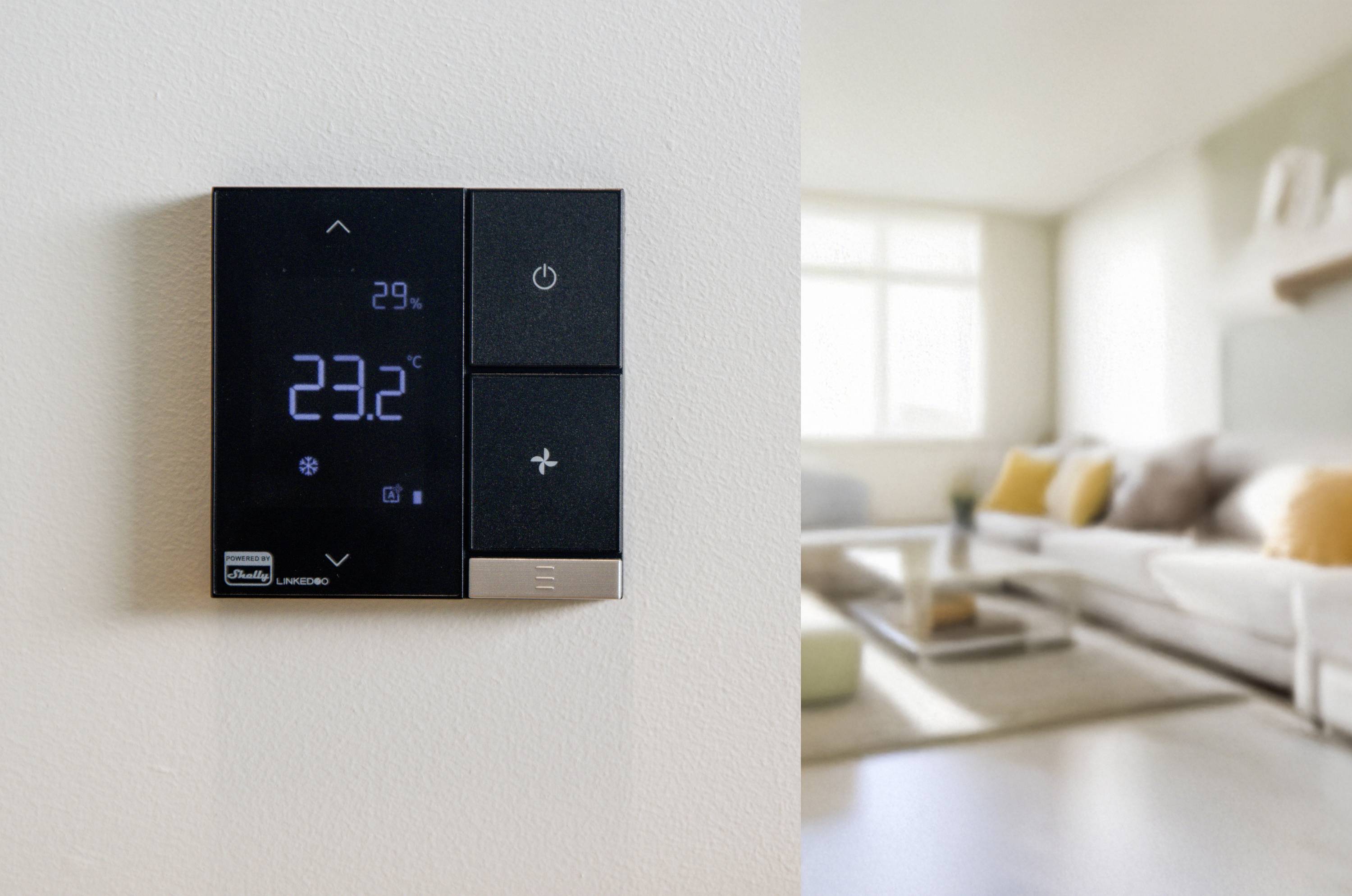 Een thermostaat aan de muur toont 23,2°C aan. Op de achtergrond is een woonkamer te zien met een bank en een tafel.