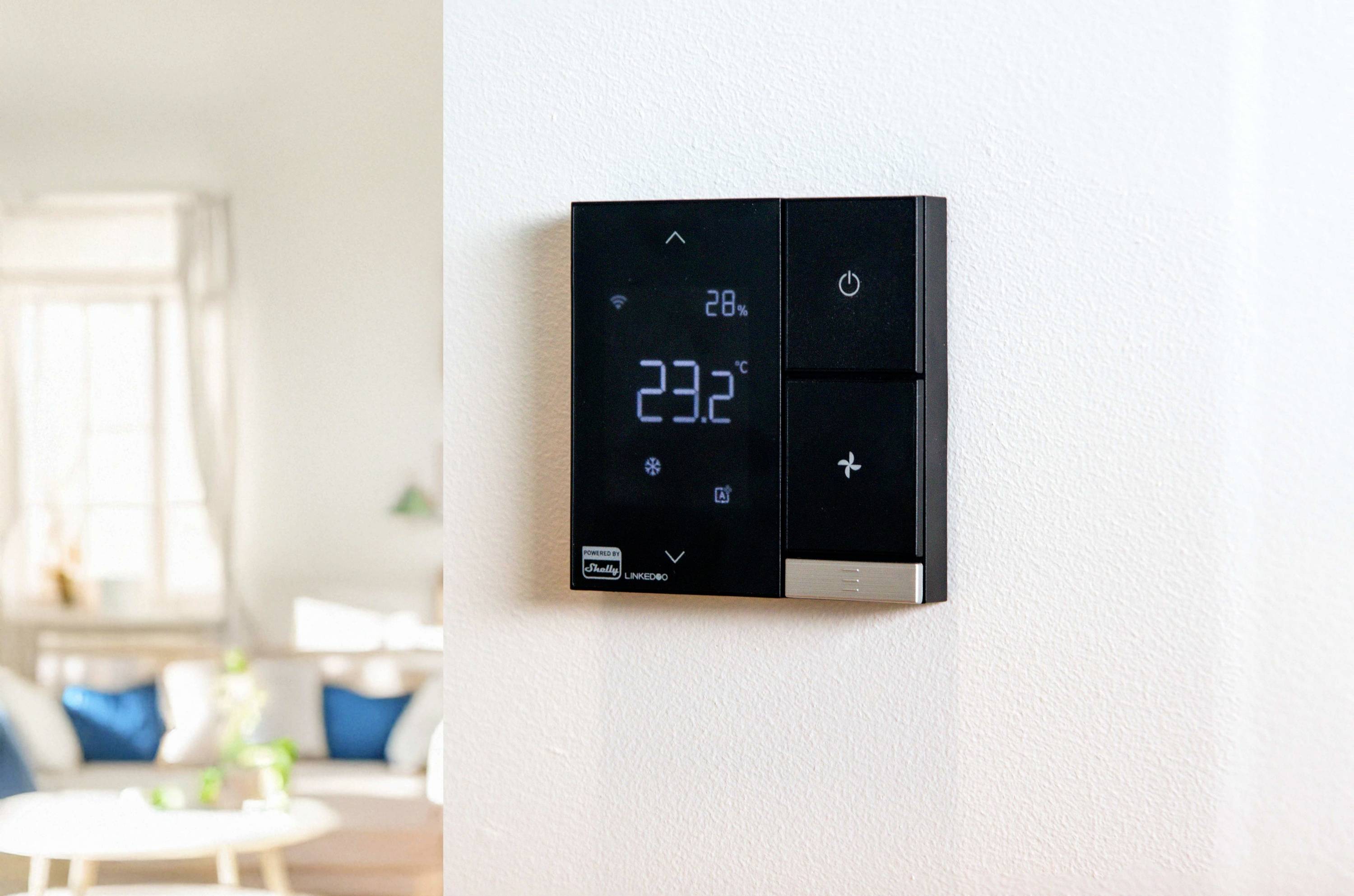 Een digitaal wandthermostaat toont '23,2°C'. De ruimte op de achtergrond is licht en modern ingericht.
