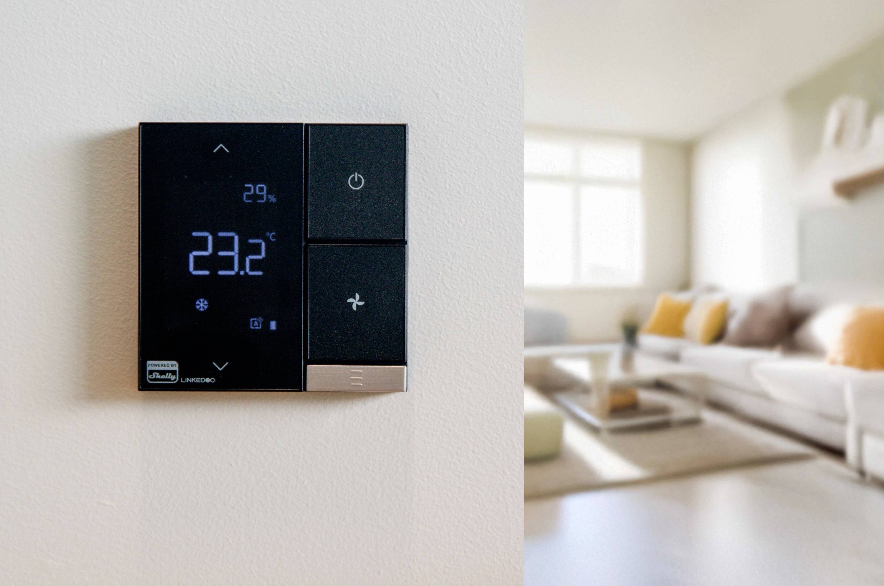 Digitale thermostaat aan de muur toont 23,2°C, woonkamer op de achtergrond met bank en kussens. Thema: moderne klimaatbeheersing.