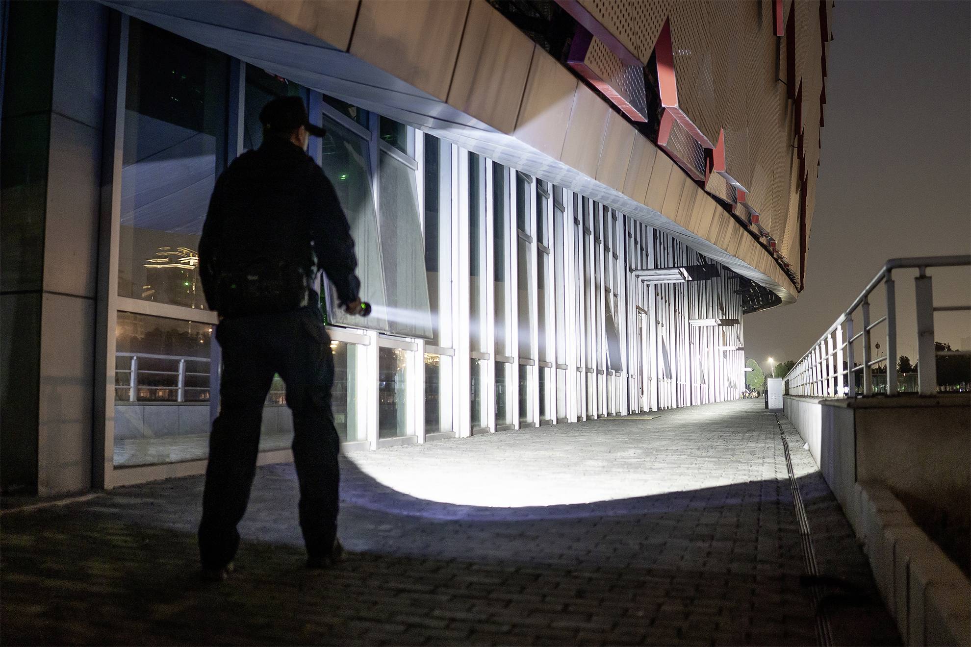 Een persoon in uniform schijnt 's nachts met een zaklamp op de grond voor een modern gebouw.