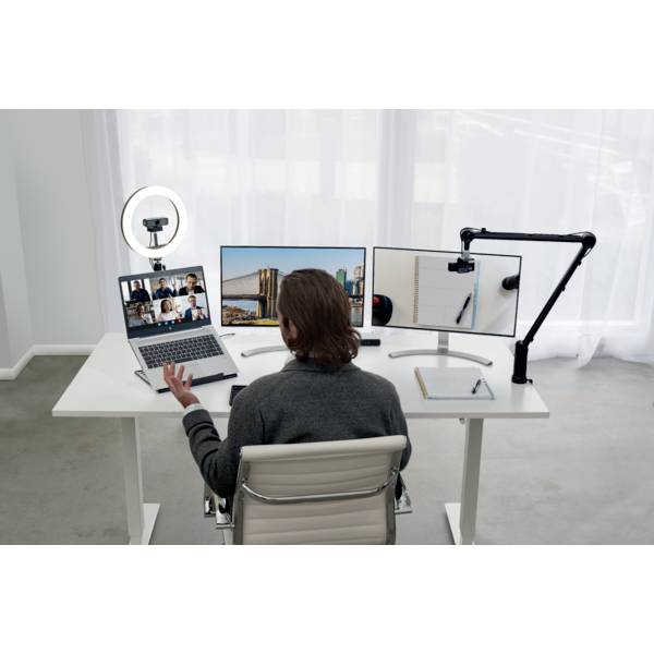 Een persoon zit aan een bureau met twee monitors, een laptop met een videovergadering en een microfoonarm, klaar voor een online vergadering.