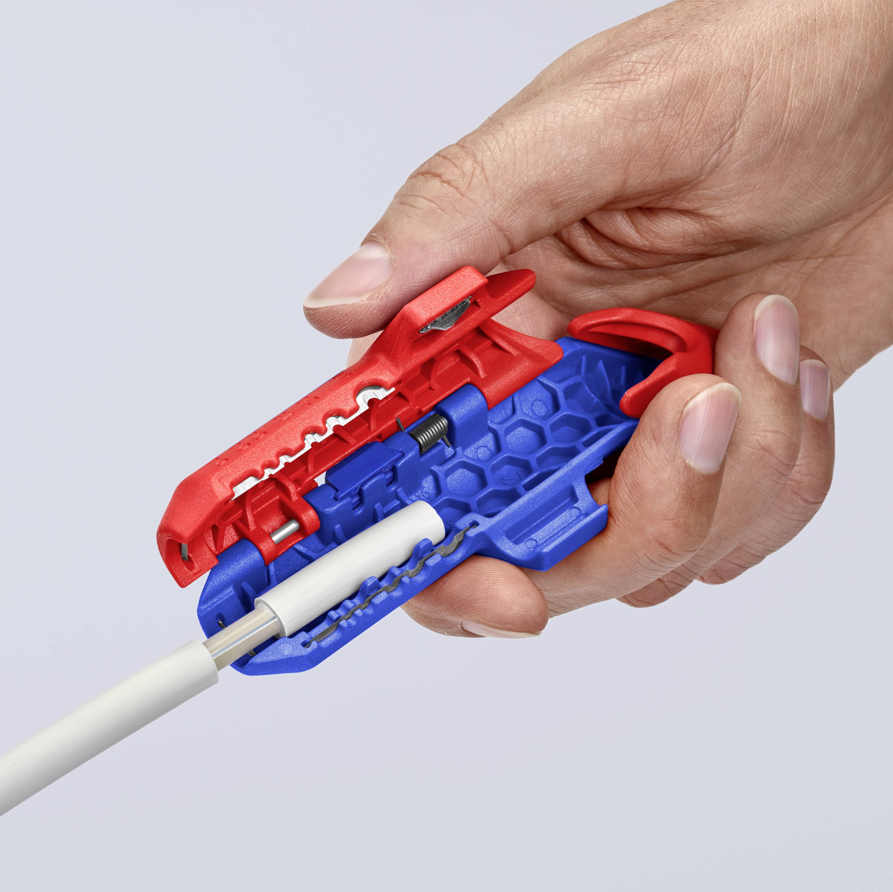 Een hand houdt een rood gereedschap vast dat een kabel ontmantelt. Het gereedschap trekt de buitenmantel weg om de binnenkabel bloot te leggen.