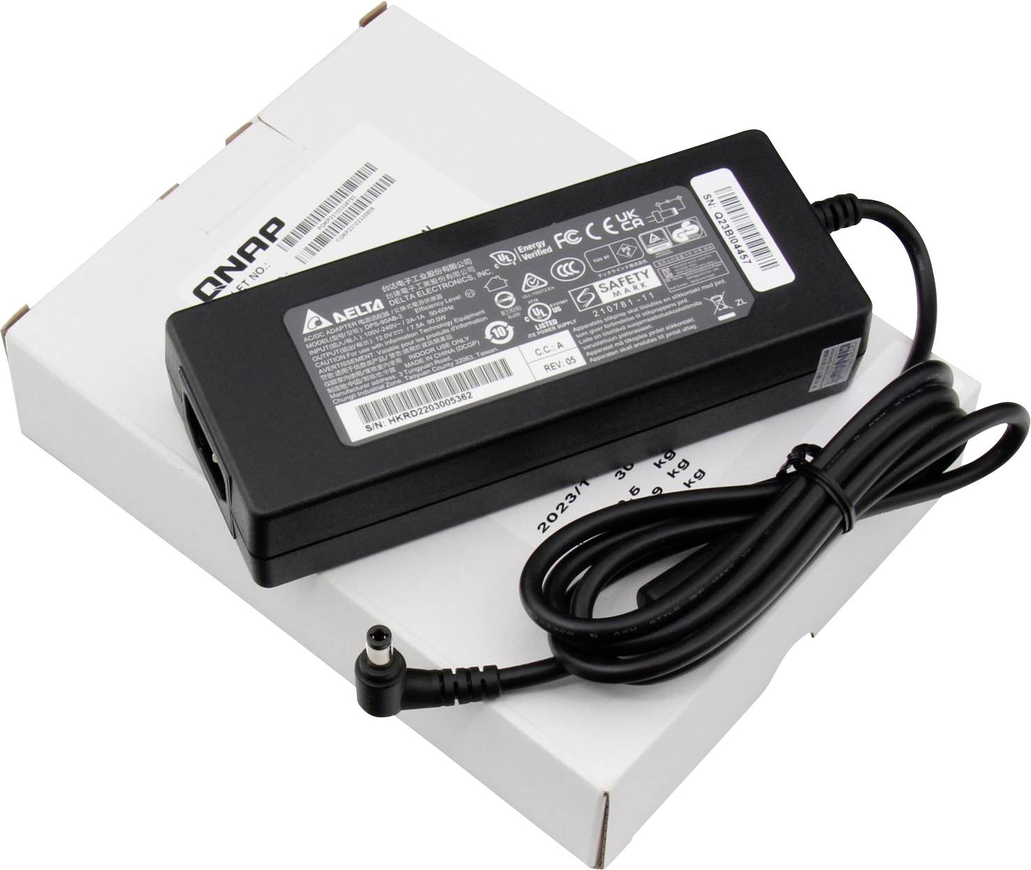 Voedingsadapter liggend op een kartonnen doos, met kabel en verschillende spanningsaanduidingen op het etiket. Geschikt voor elektronische apparaten.