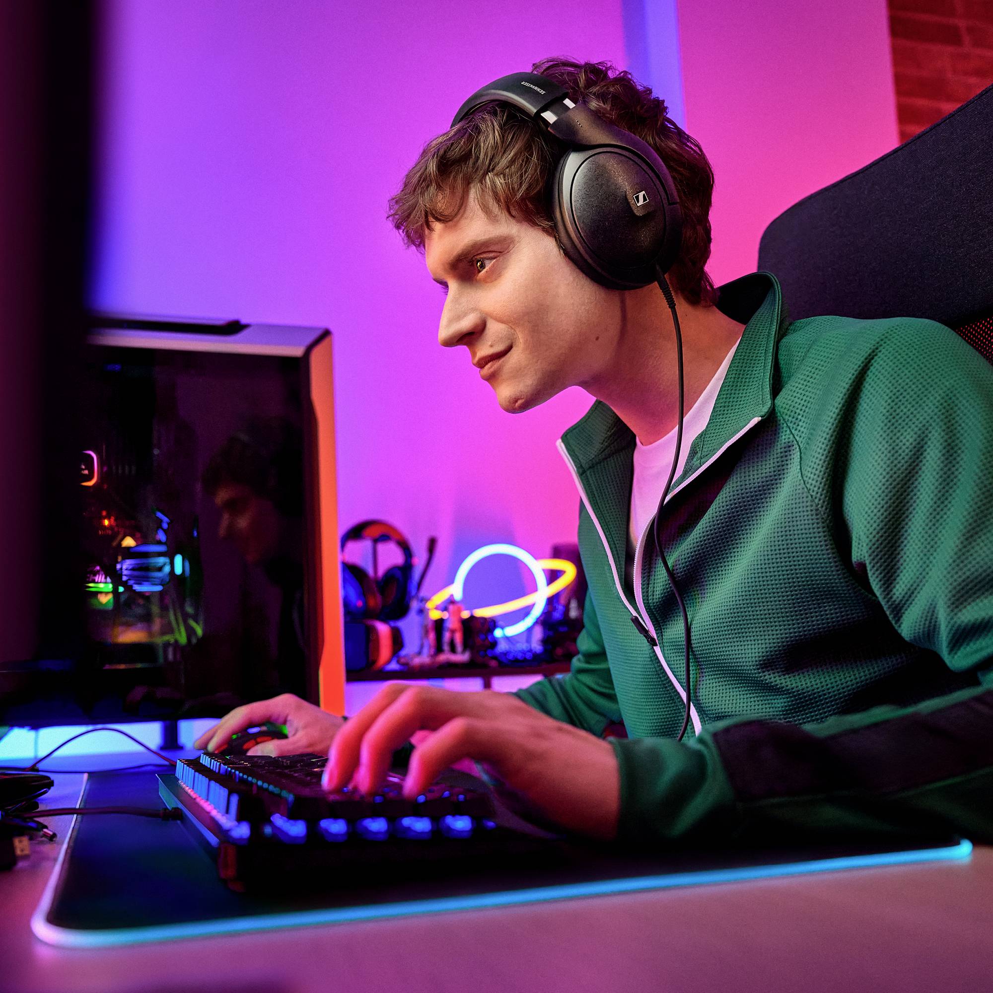 Een persoon speelt op de computer, draagt koptelefoons en een groene jas. De ruimte is kleurrijk verlicht.