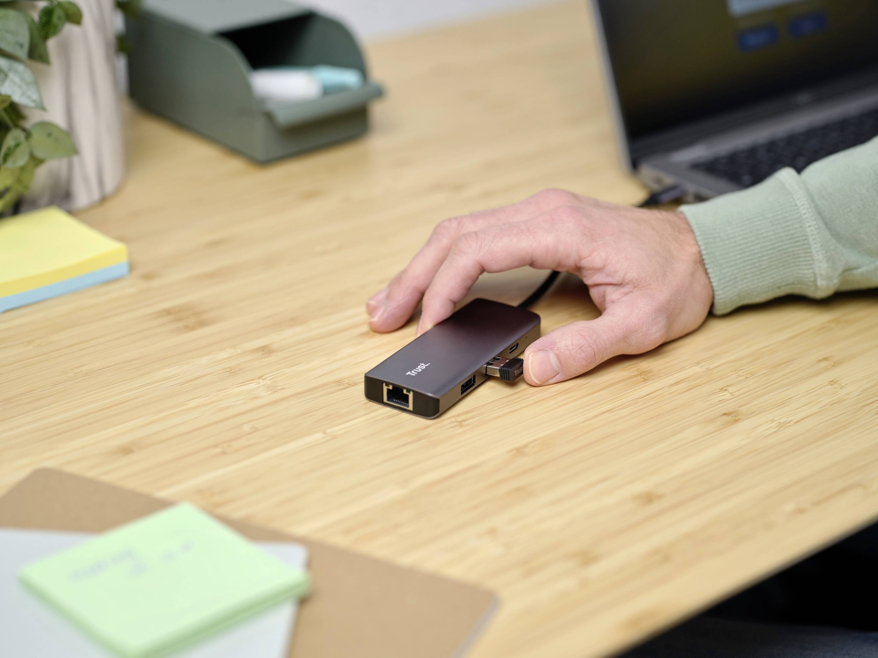Een hand raakt een zwarte USB-C-hub aan op een houten tafel, terwijl naast hem een laptop is opengevouwen.