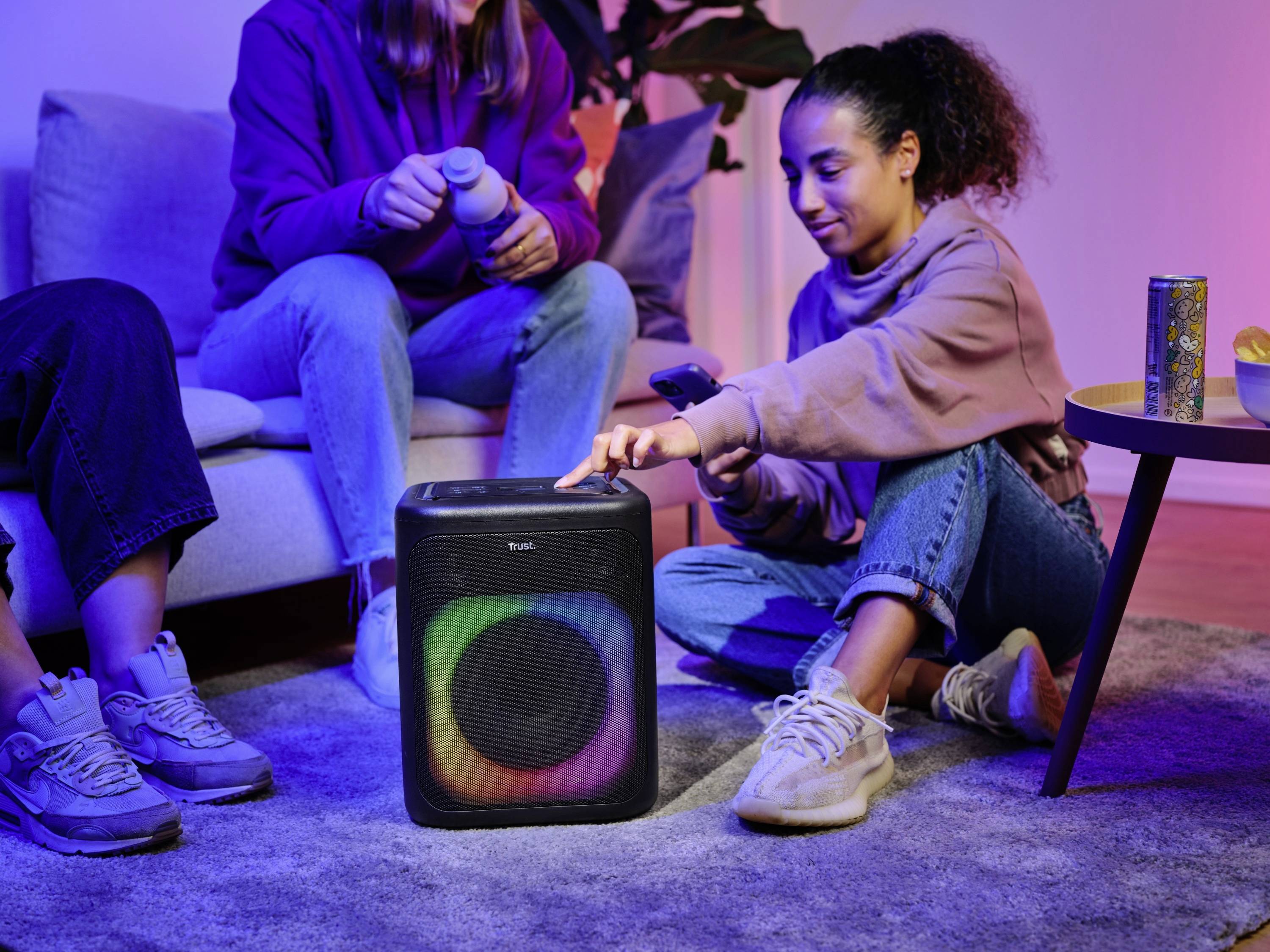Twee personen zitten in de woonkamer. Één persoon bedient via de smartphone een draagbare luidspreker met gekleurde verlichting.