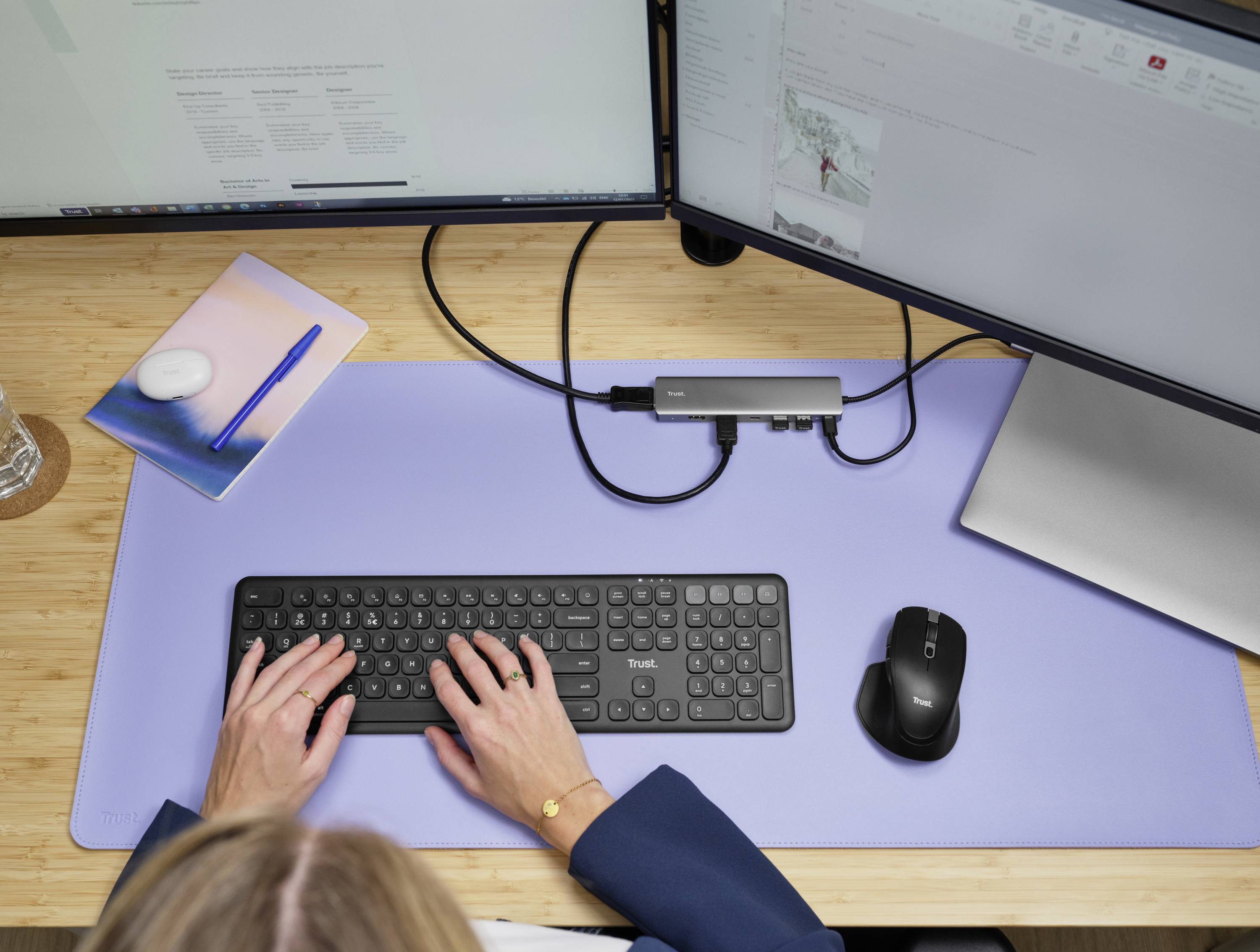 Een persoon werkt aan een bureau met twee monitors, een toetsenbord en een muis. Het bureau is bedekt met een lichtblauwe onderlegger.