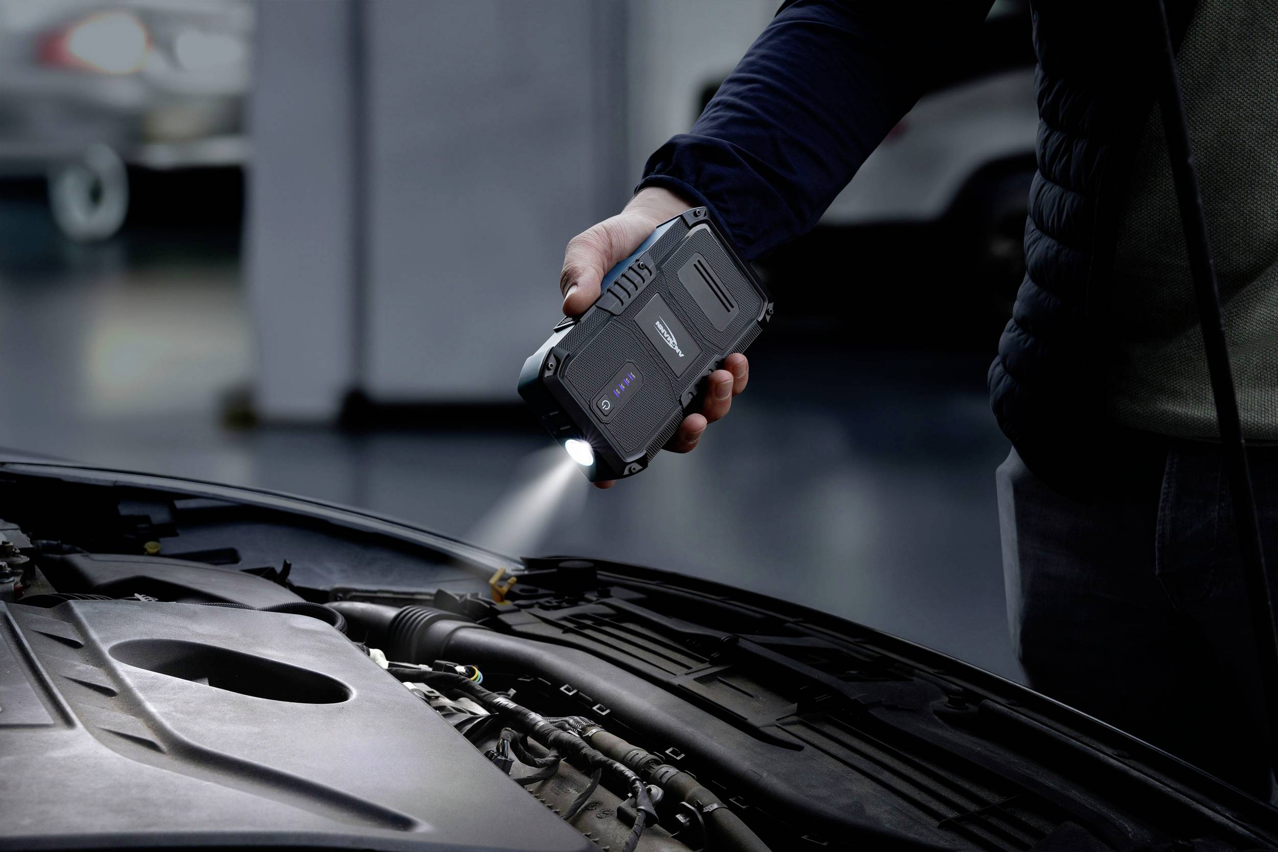 Een persoon houdt een diagnoseapparaat boven een open automotor in het interieur van een werkplaats. Het apparaat verlicht het motorgedeelte.