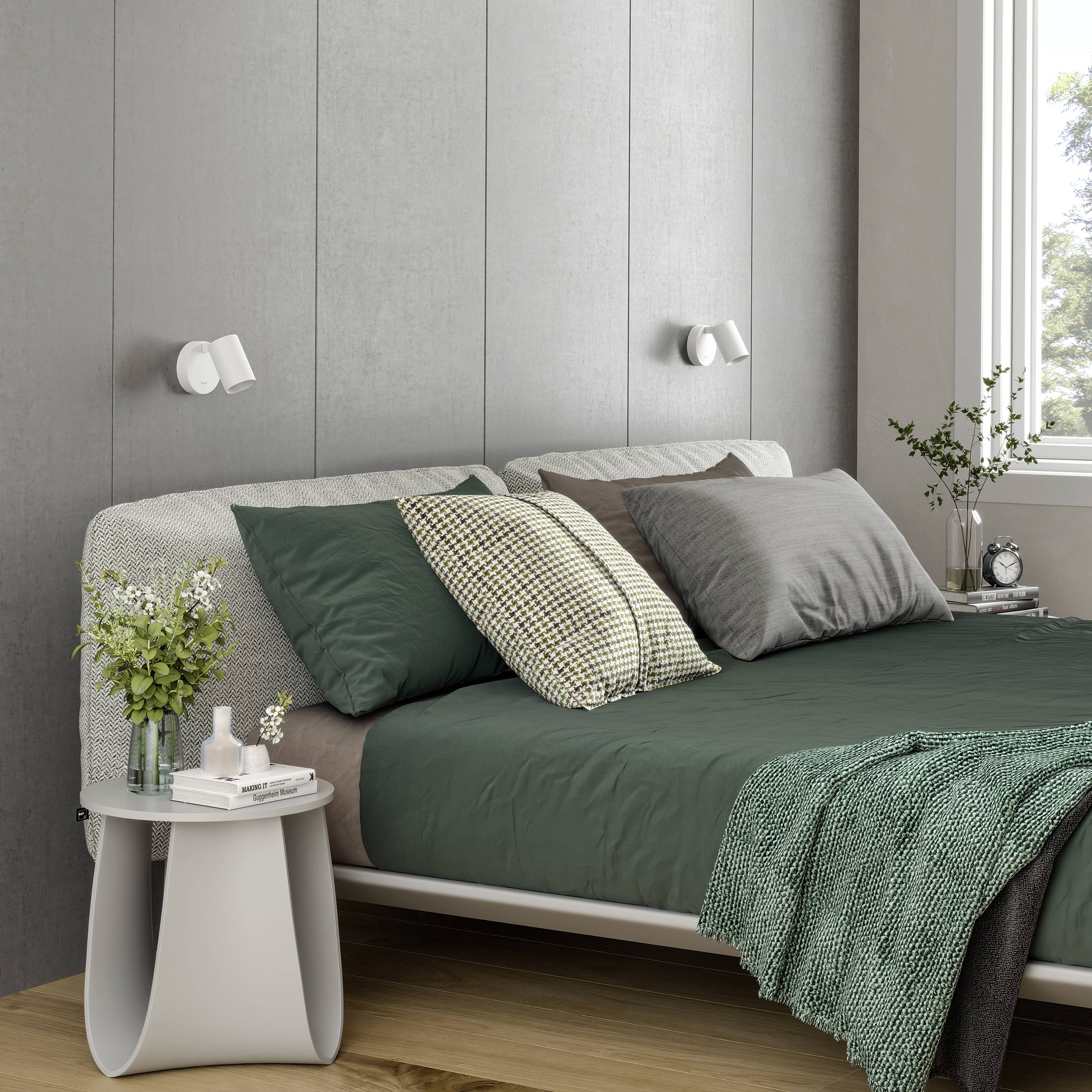 Gezellige slaapkamer met een modern grijs bed, groene dekens en kussens. Nachtkastje met planten en een lamp, daglicht door het raam.