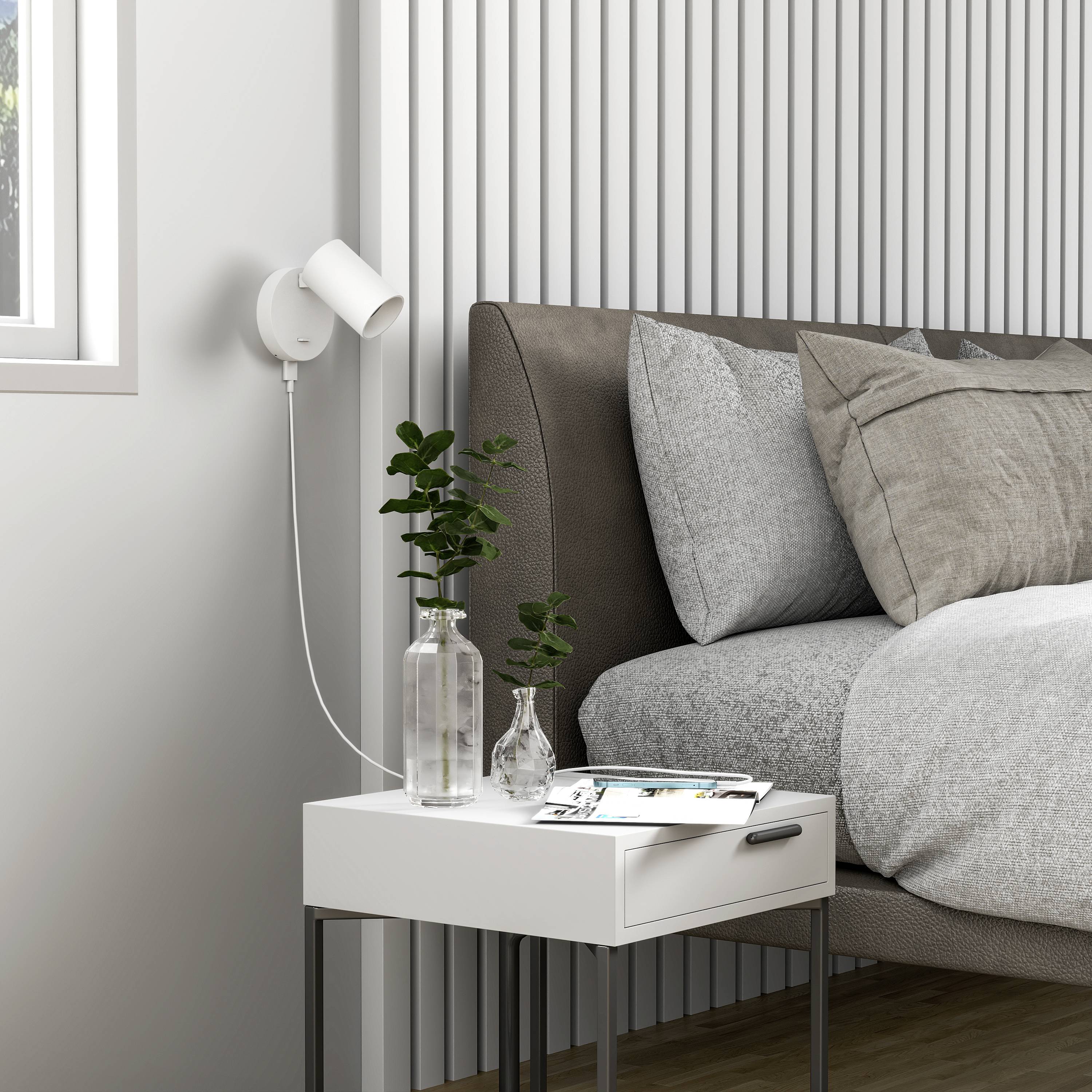 Bed met grijze beddengoed, drie kussens, nachtkastje met vaas, plant, boek en tafellamp in een moderne, lichte kamer.