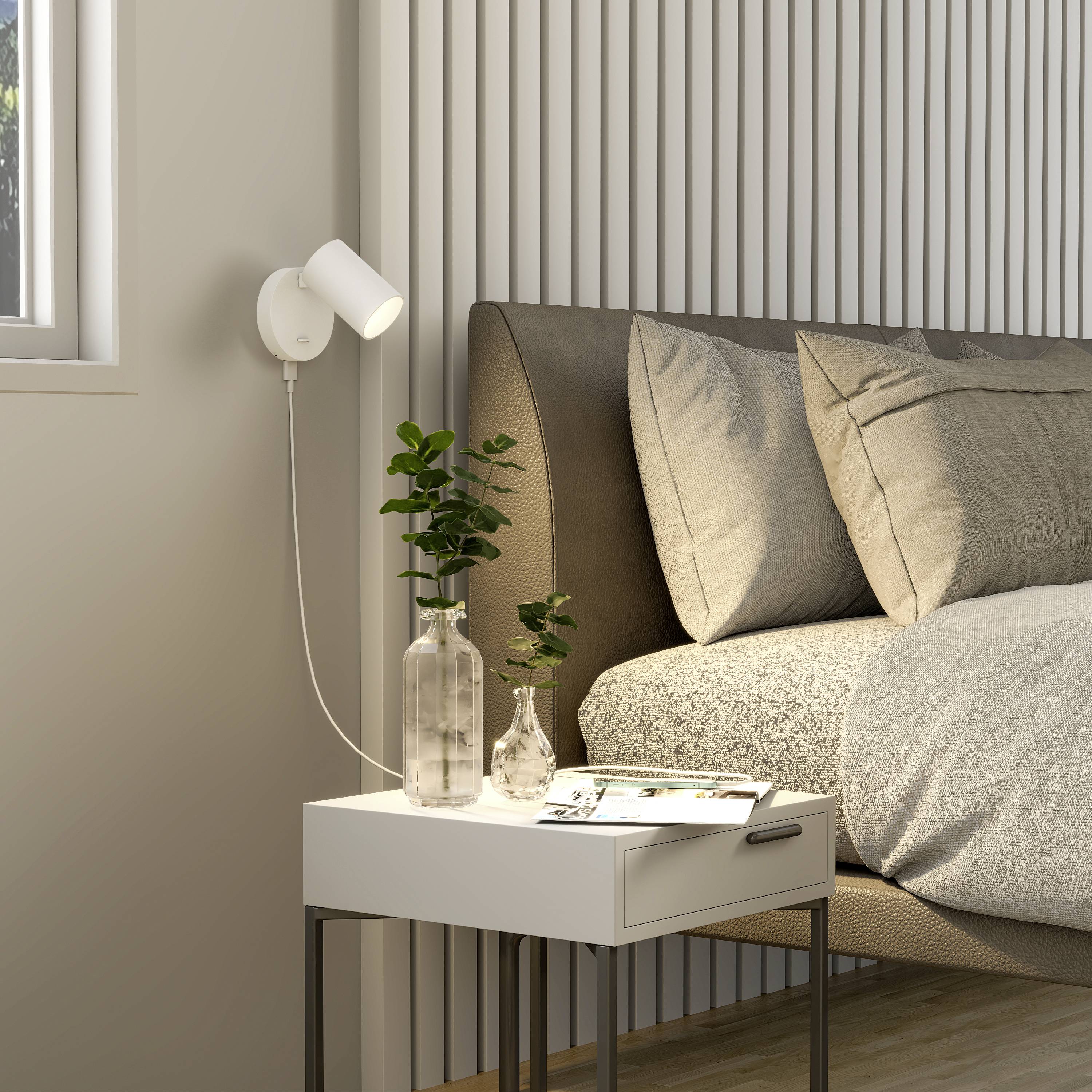 Een modern slaapkamer met een nachtkastje waarop een plant en boeken staan. Een bed met grijze kussens en houten bekleding.