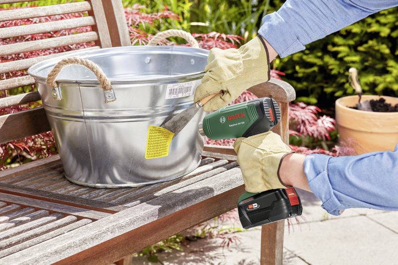 Een persoon verwijdert met een Bosch-accu-schraper een etiket van een zilveren emmer die op een houten bank in de tuin staat.