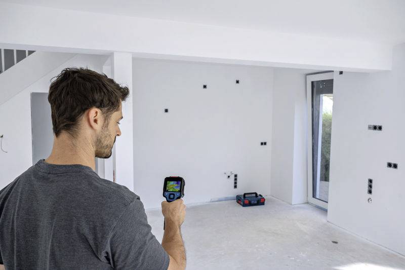 Een persoon gebruikt een warmtebeeldcamera om een muur in een lege ruimte te inspecteren. De vloer is ruw, zonder vloerbedekking.