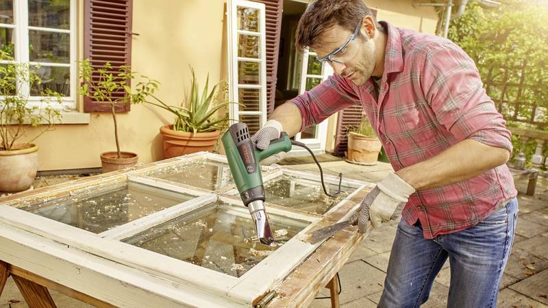 Een man repareert een vensterkozijn met een heteluchtpistool buiten. Hij draagt een veiligheidsbril en handschoenen.