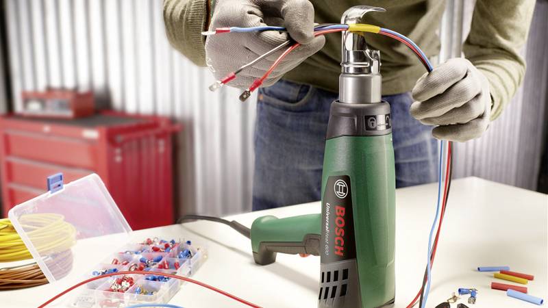 Een persoon houdt een groen Bosch-verwarmingstoestel vast en werkt met kabels aan een werkbank. De omgeving toont gereedschappen en reserveonderdelen.