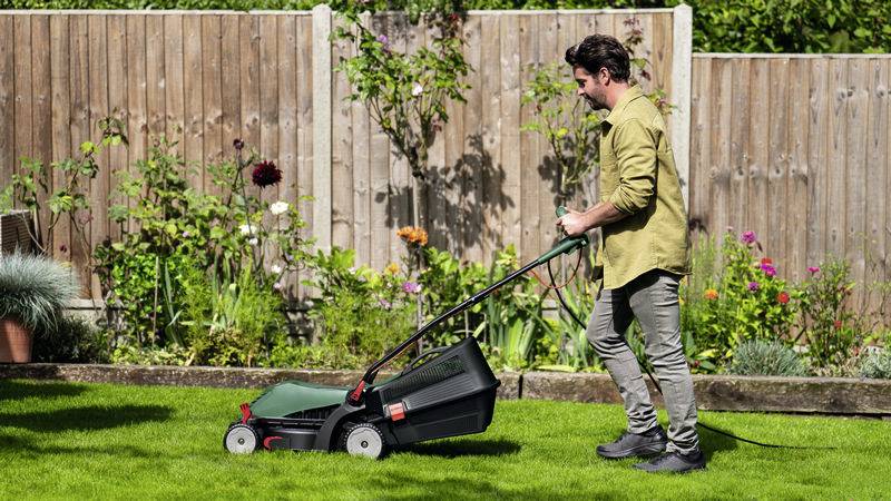 Een persoon maait het gazon met een grasmaaier in een tuin. Op de achtergrond zijn bloemen en een houten hek zichtbaar.
