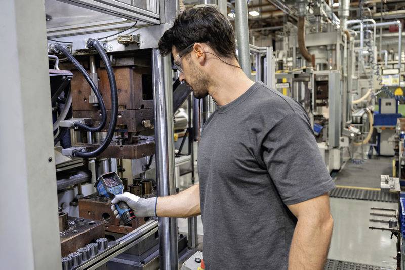 Een man in een grijs T-shirt en witte handschoen controleert productiemachines met een apparaat in een industriële omgeving.