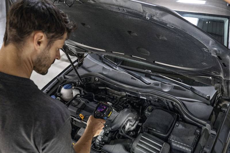 Een man staat met een diagnose-apparaat voor een open auto. De motorruimte is zichtbaar, hij controleert technische gegevens.
