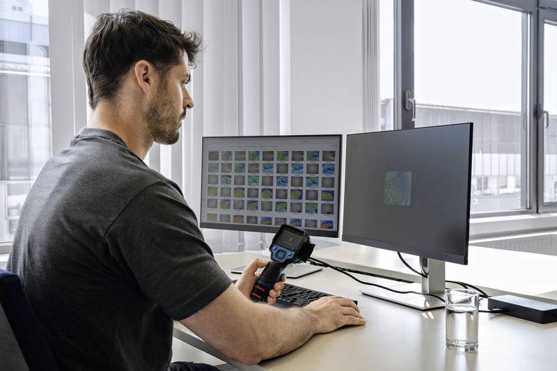 Een persoon zit aan een bureau en gebruikt een apparaat voor schermkalibratie op een monitor. Op de achtergrond zijn twee beeldschermen zichtbaar.