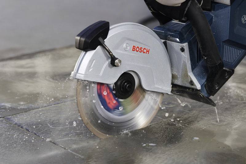 Een elektrische cirkelzaag snijdt een stenen plaat, terwijl water wordt gespetterd om de zaagblad te koelen. De naam 'Bosch' is zichtbaar.