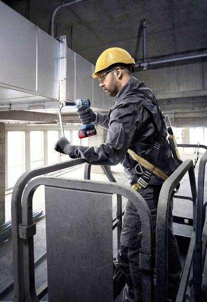 Een bouwvakker met een veiligheidshelm en veiligheidsgordel staat op een hoogwerker en gebruikt een elektrisch gereedschap om leidingen te installeren.