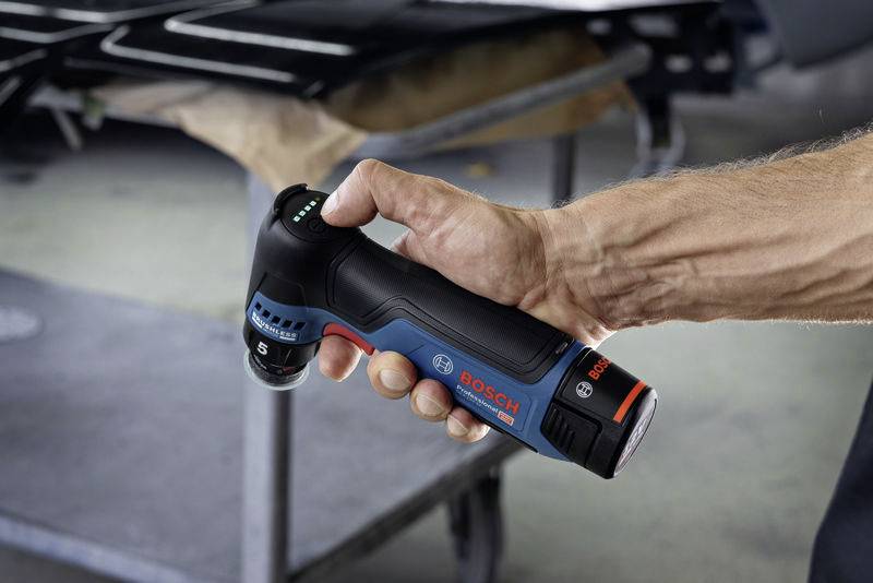 Een hand houdt een blauwe en zwarte Bosch-accuschroevendraaier met een rood logo; het gereedschap wordt gebruikt in een werkplaats.