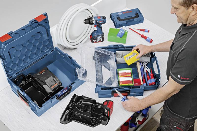 Een man werkt aan een tafel met geopende gereedschapskoffers, een accuboormachine en elektriciteitskabels. Gereedschappen en toebehoren liggen uitgespreid.