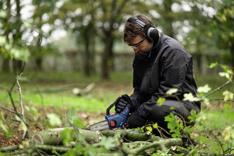 Een man in beschermende uitrusting knielt in het bos en zaagt met een kettingzaag een omgevallen boomstam. Bladeren en takken liggen verspreid rond hem heen.