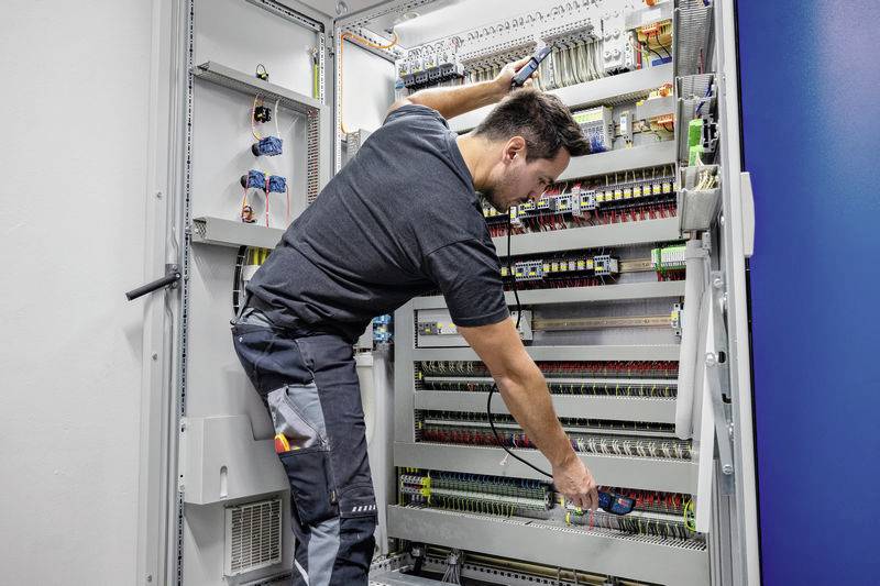 Een technicus werkt aan een schakelkast, controleert bedrading en verbindingen. De kast bevat talrijke elektronische componenten en leidingen.