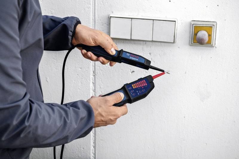 Een persoon controleert de spanning aan een stopcontact met een elektrisch meetapparaat. Het apparaat toont een LED-display.