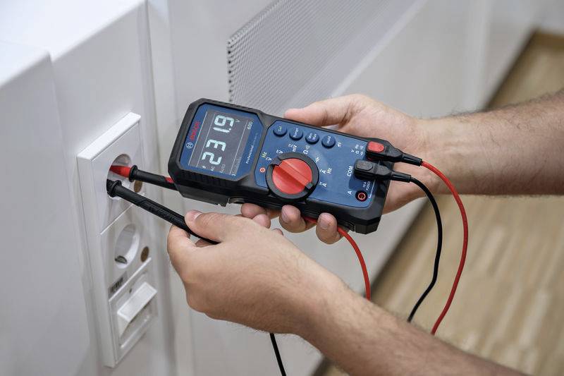 Persoon meet spanning met een digitaal multimeter bij een stopcontact. Display toont meetwaarden.