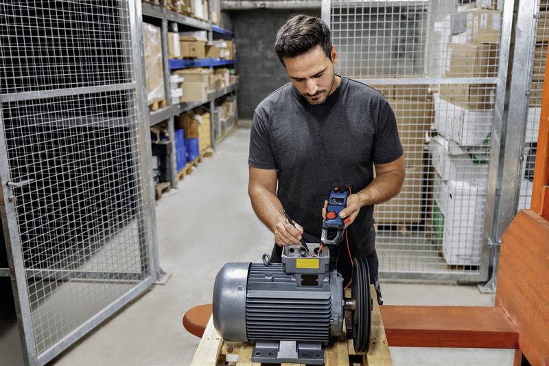 Een man meet met een multimeter aan een grote elektromotor in een magazijn. Op de achtergrond staan stellingen met dozen.