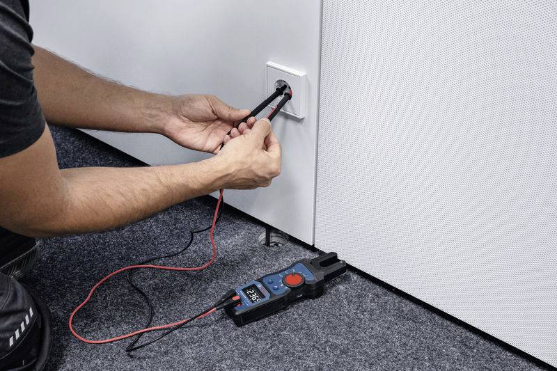 Een persoon controleert de spanning van een stopcontact met een multimeter. Platte hoek op een grijze tapijtvloer.