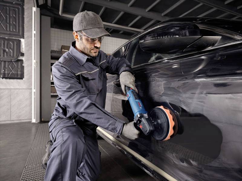Een monteur polijst de lak van een zwarte auto met een polijstmachine in een werkplaatsomgeving.