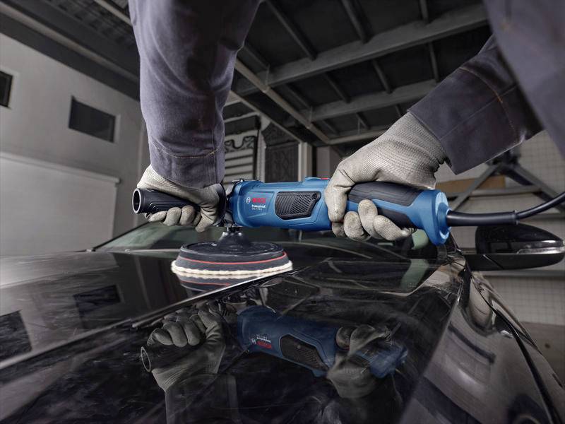 Een persoon polijst een zwarte auto met een blauwe polijstmachine in een garage. Handen dragen handschoenen en houden het apparaat stevig vast.